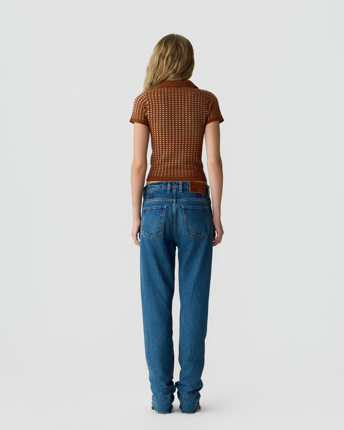 Rear view of a woman wearing a textured short-sleeve brown top and high-waisted blue jeans, standing against a plain light gray background.