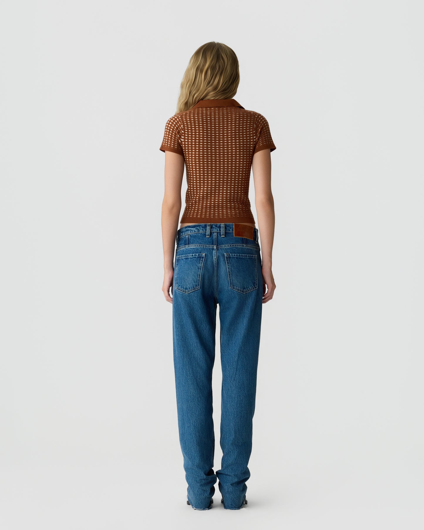 Rear view of a woman wearing a textured short-sleeve brown top and high-waisted blue jeans, standing against a plain light gray background.