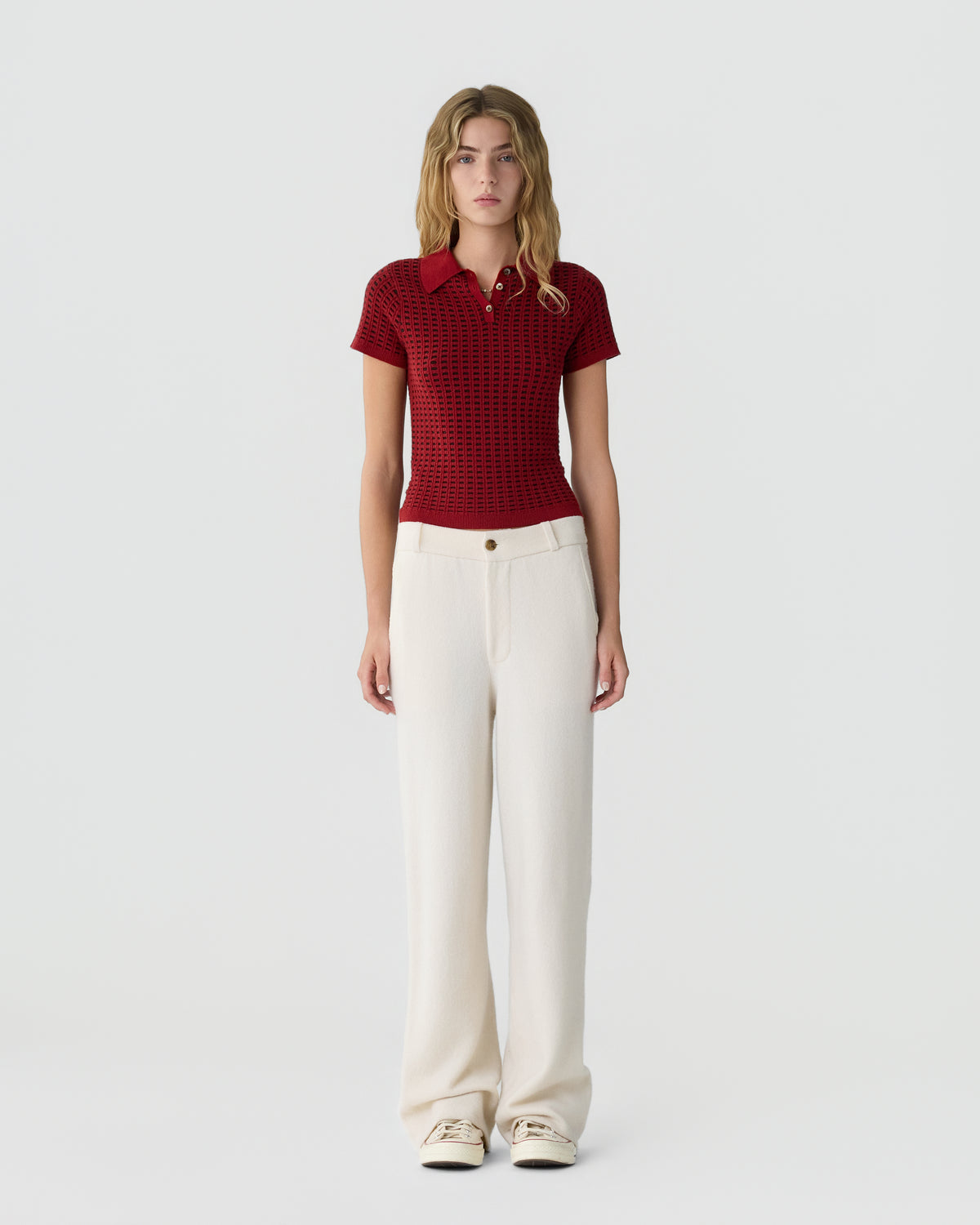 A young woman stands against a plain background wearing a short-sleeved, red textured polo shirt paired with high-waisted, cream wide-leg pants and light-colored sneakers.