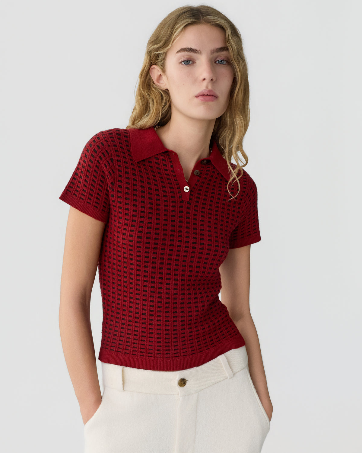 Young woman wearing a short-sleeved, red textured knit polo shirt paired with high-waisted white pants, standing against a plain light gray background with hands in pockets.