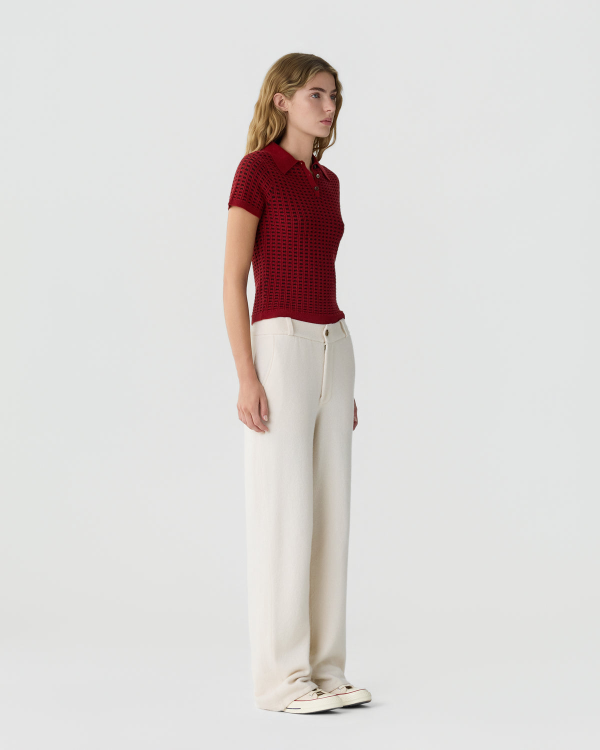 Profile view of a woman wearing a red short-sleeve knit polo shirt paired with high-waisted, wide-leg cream trousers and light-colored sneakers against a plain background.