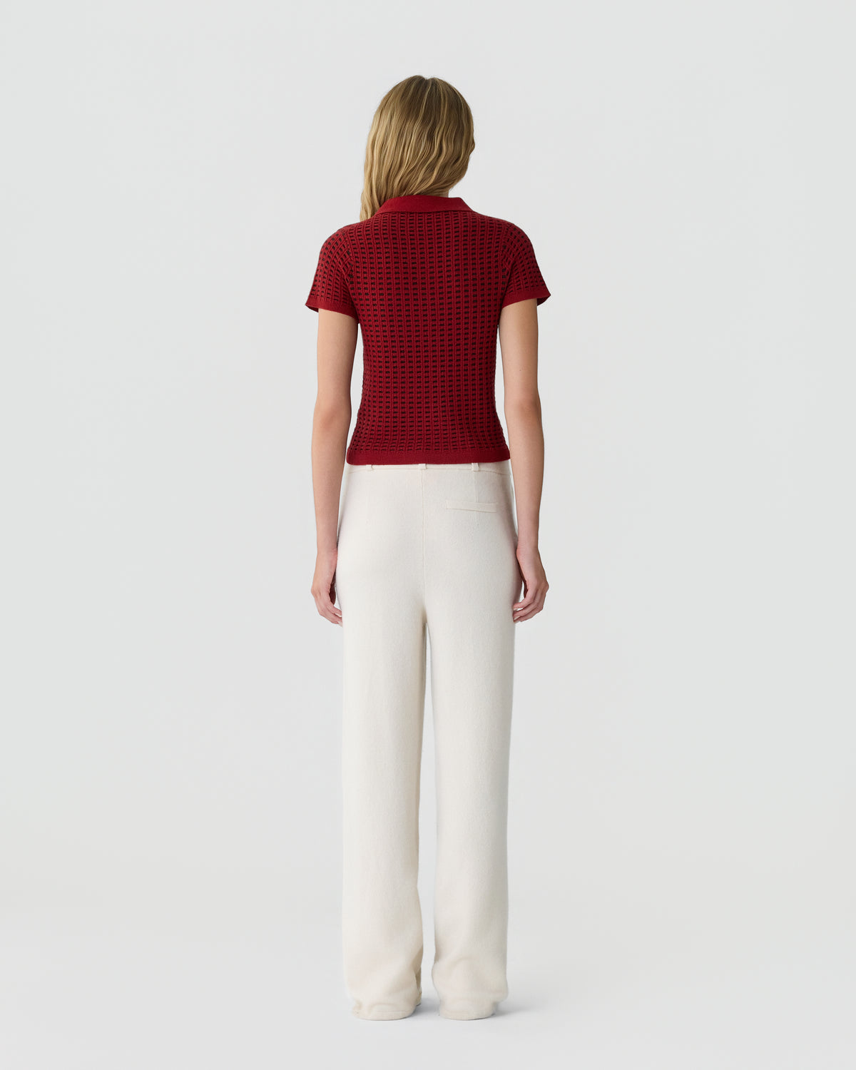 Woman with long blonde hair wearing a short-sleeve red textured top and high-waisted wide-leg white pants, standing facing away against a plain background.