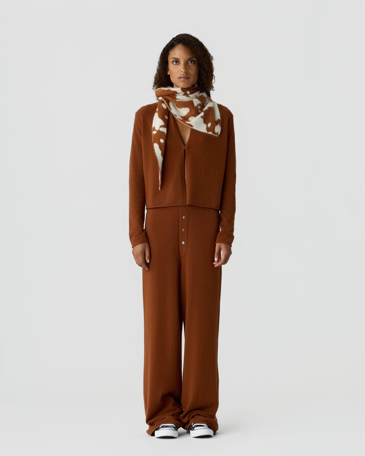Woman wearing a brown knit top and wide leg trousers with a patterned scarf and white sneakers against a light gray studio background