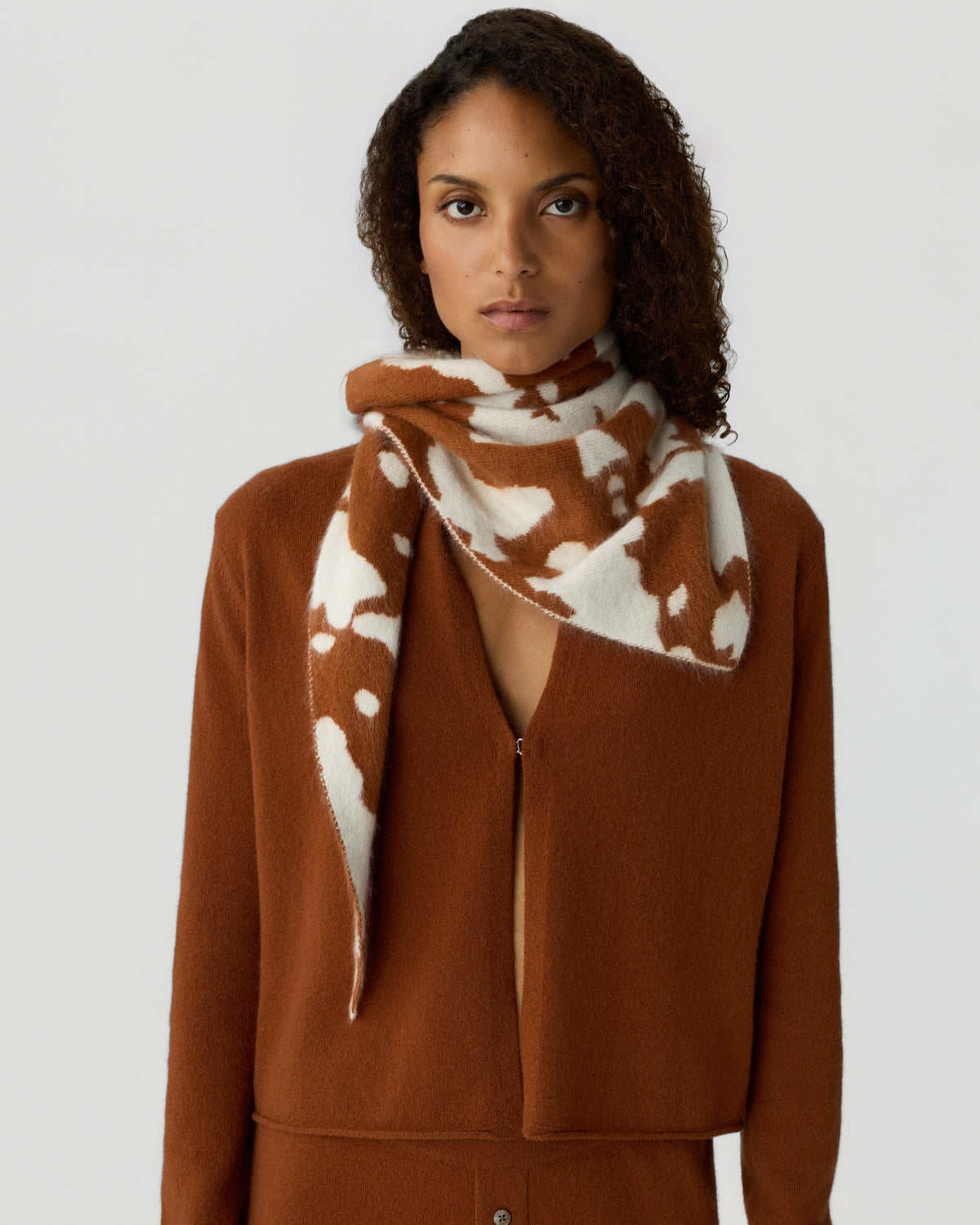 Model wearing a rust brown knit cardigan with a cream scarf featuring an abstract brown pattern against a light gray background