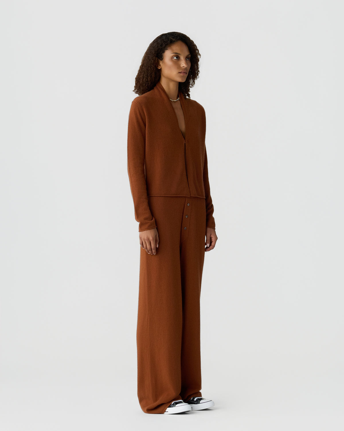 Woman standing against a light gray background wearing a brown cashmere cardigan with matching wide leg trousers and white sneakers