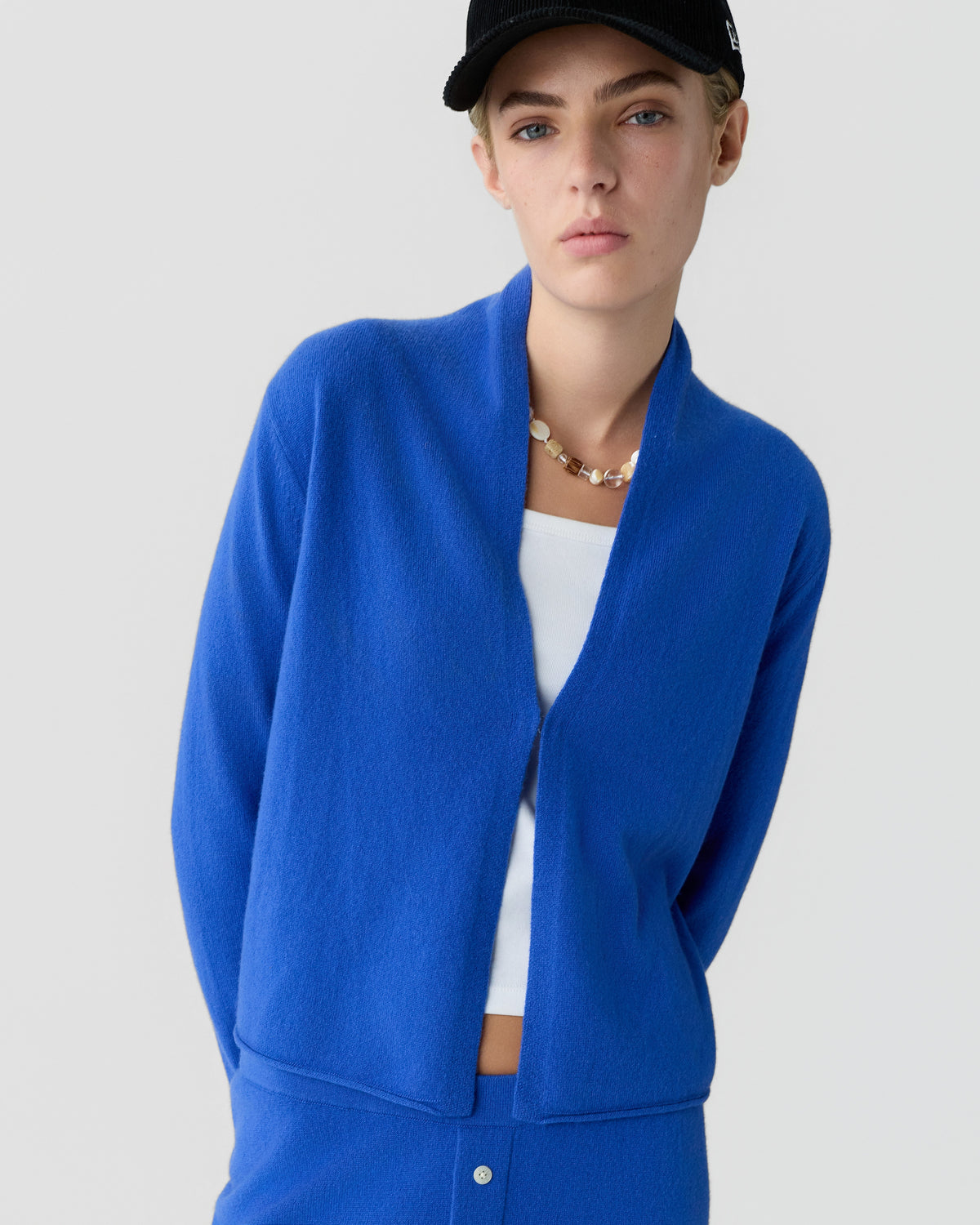 Model wearing a bright blue open-front cardigan over a white top with a black cap and necklace against a light gray background