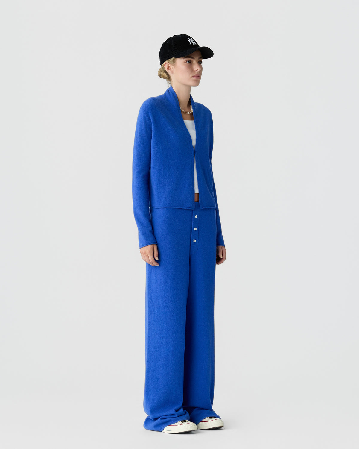 Woman wearing a bright blue cardigan and matching wide leg trousers with a black baseball cap and white sneakers against a light gray studio background