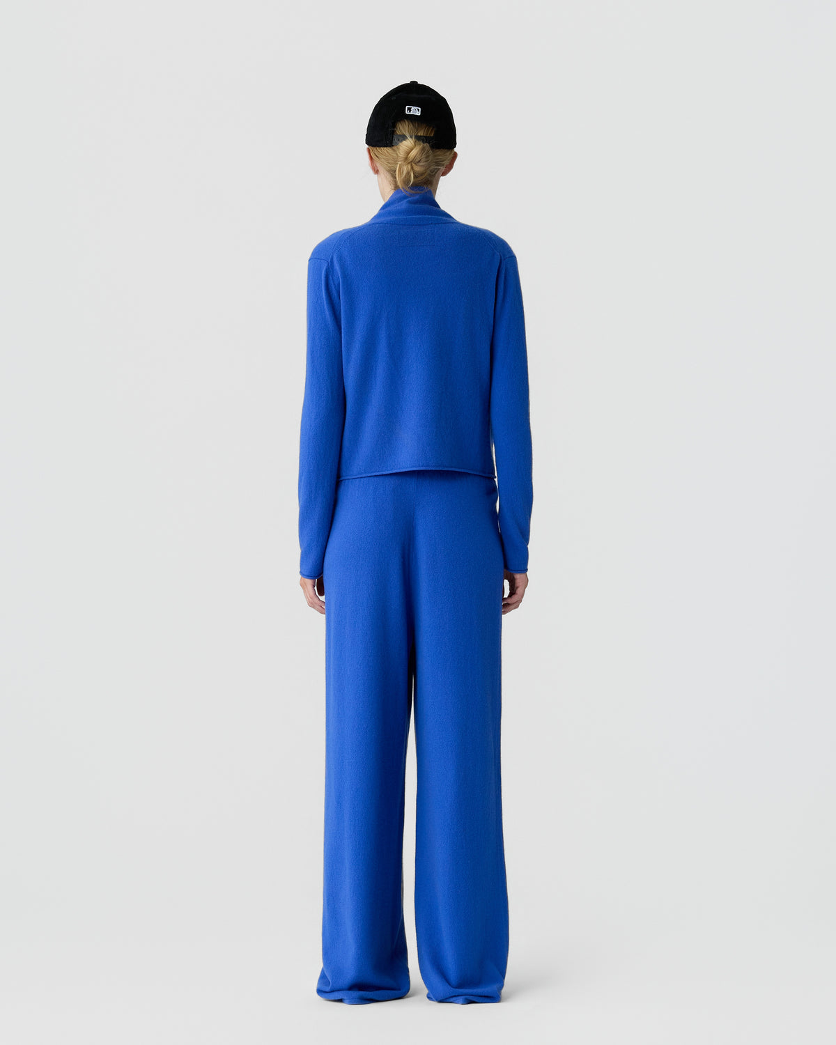 Rear view of a person in a bright blue cashmere sweater and matching wide leg pants with a black cap against a light gray background