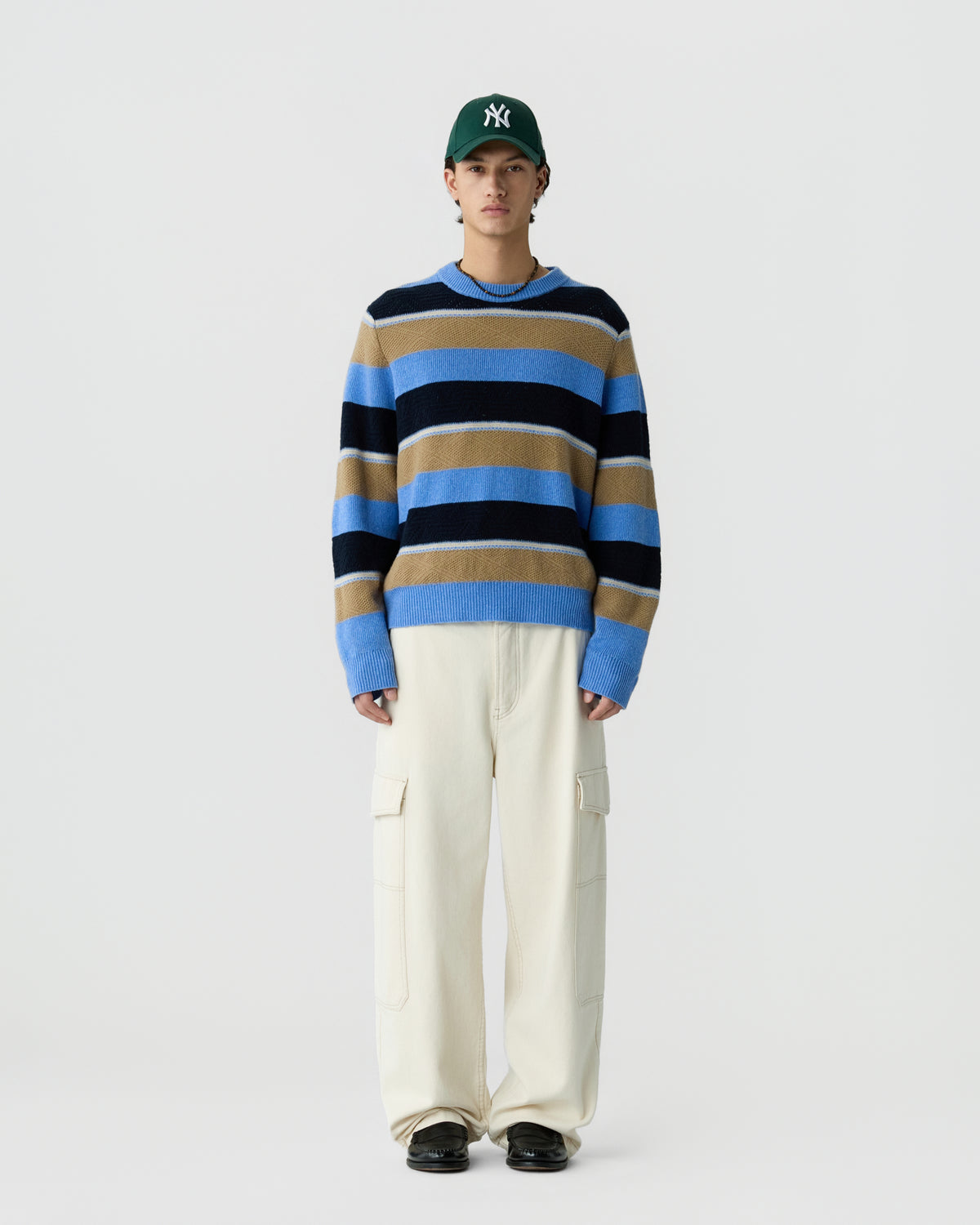 Young man wearing a green baseball cap, a blue, tan, and black horizontally striped sweater, loose white cargo pants, and black shoes, standing against a plain light background.