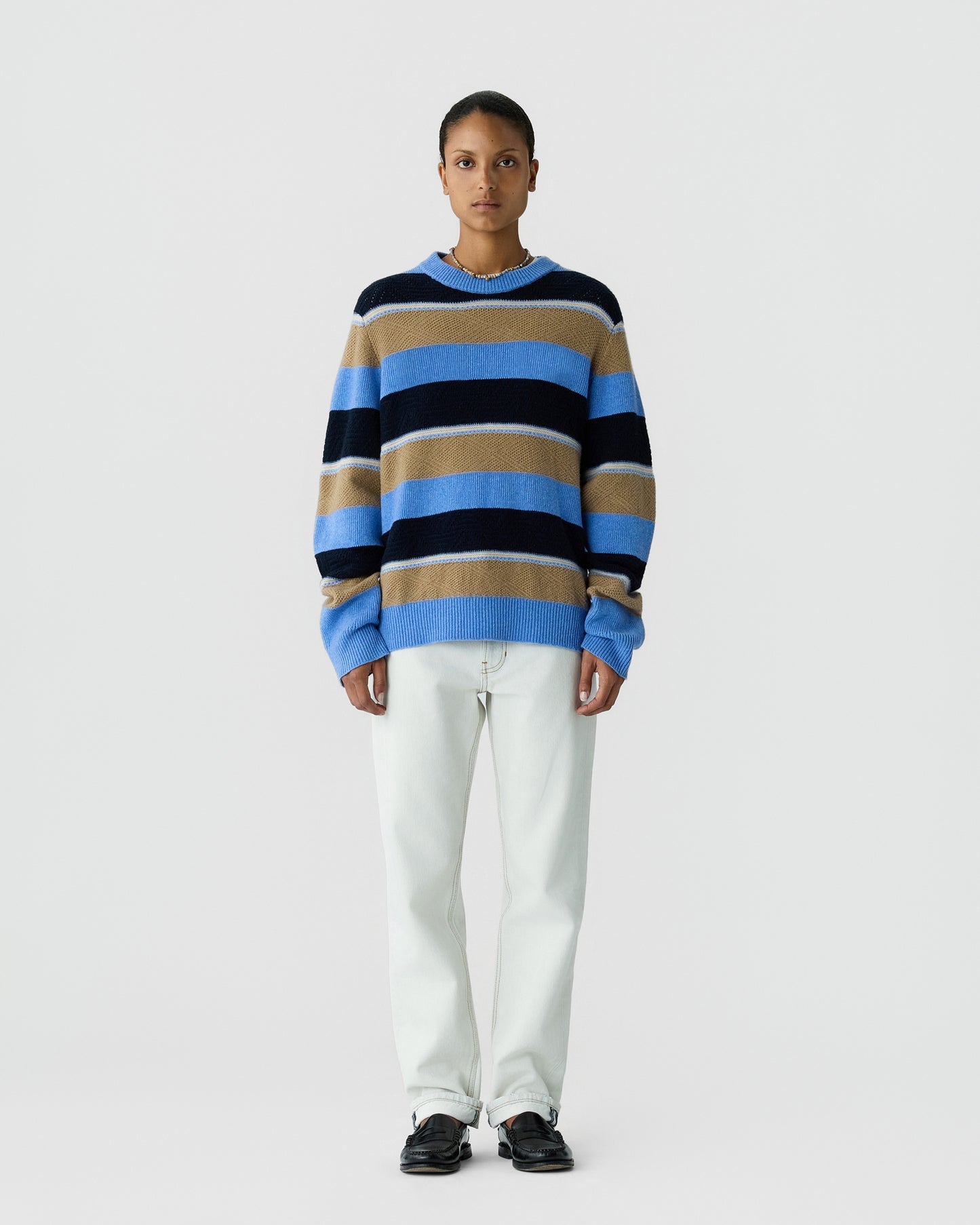 Person standing against a plain background wearing a loose striped sweater in blue, black, and tan paired with off-white pants and black shoes.