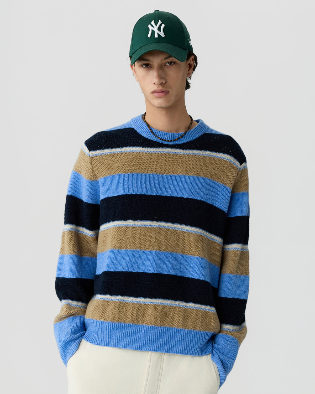 Young man wearing a green baseball cap and a knitted sweater with wide horizontal stripes in blue, black, and tan, paired with light-colored pants, standing against a plain background.