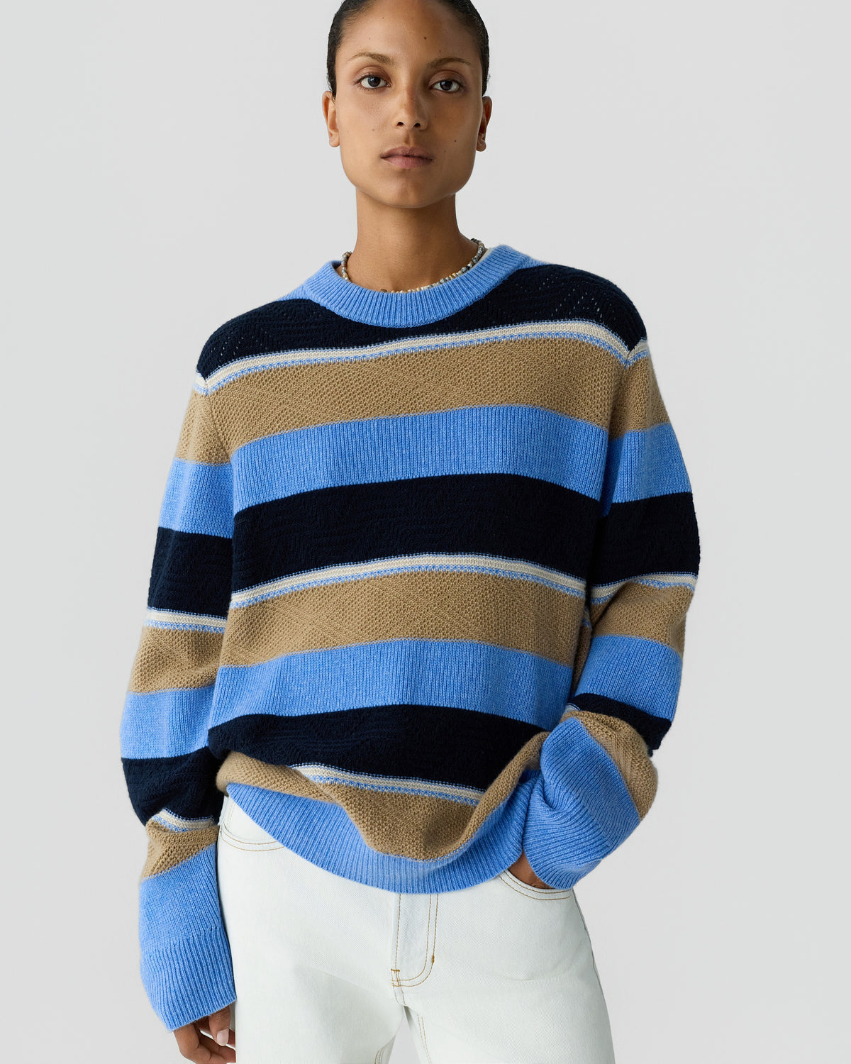 Person wearing a long-sleeve sweater with wide horizontal stripes in light blue, navy, and beige, paired with white pants against a plain light gray background.
