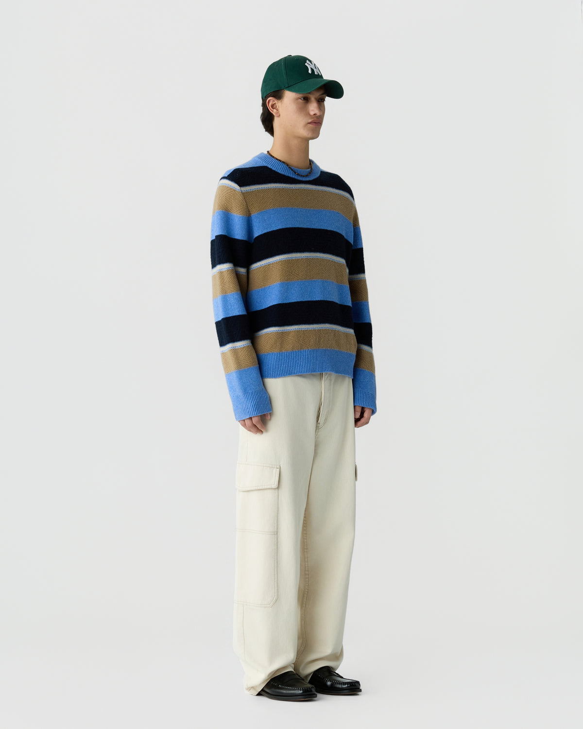 Young man wearing a green baseball cap, a long-sleeve striped sweater in blue, black, and tan colors, cream-colored wide-leg pants with pockets, and black shoes, standing against a white background.