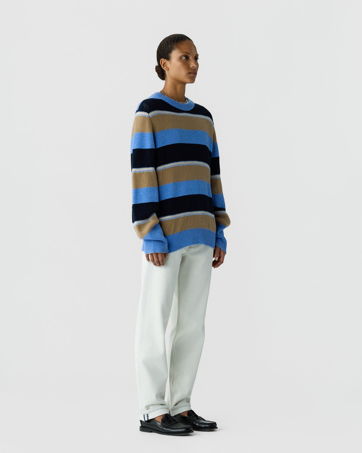 Woman standing against a plain background wearing a loose-fitting sweater with horizontal stripes in blue, navy, beige, and white, paired with white pants and black loafers.