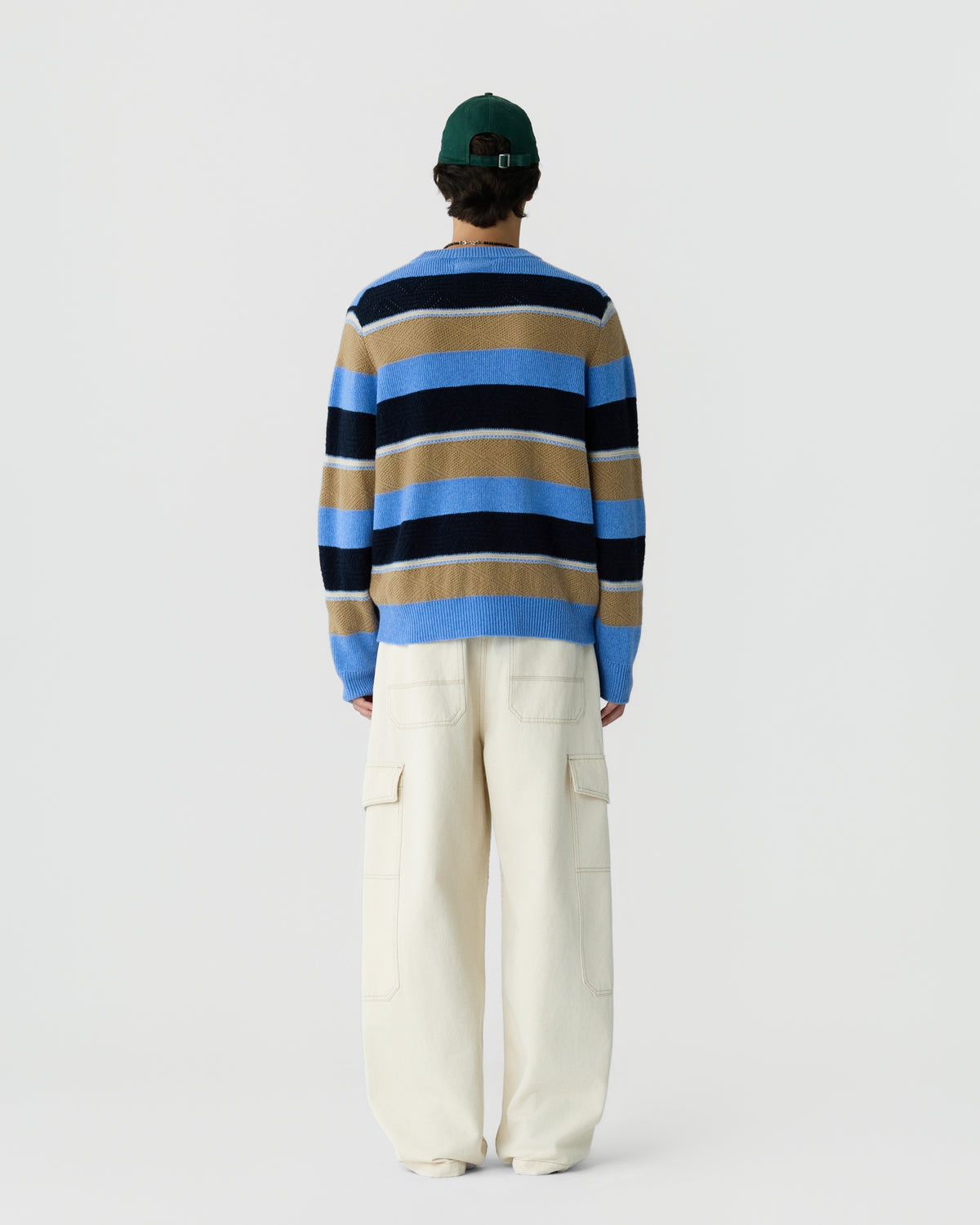 Person wearing a blue, black, and tan horizontal striped sweater with cream-colored wide cargo pants and a dark green cap, viewed from the back against a plain background.