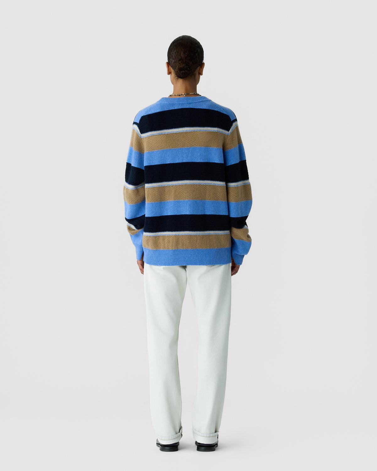 Person standing facing away wearing a long-sleeve crewneck sweater with horizontal stripes in blue, beige, and black, paired with white pants and black shoes against a plain light background.