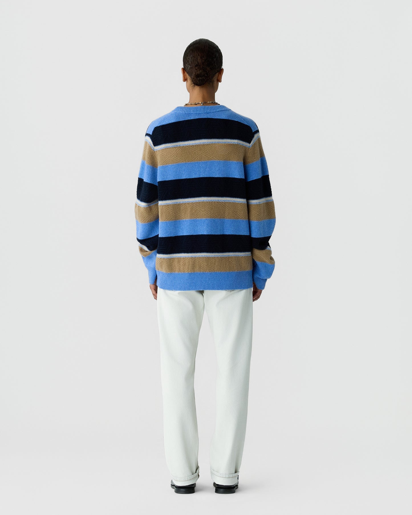 Person standing facing away wearing a long-sleeve crewneck sweater with horizontal stripes in blue, beige, and black, paired with white pants and black shoes against a plain light background.