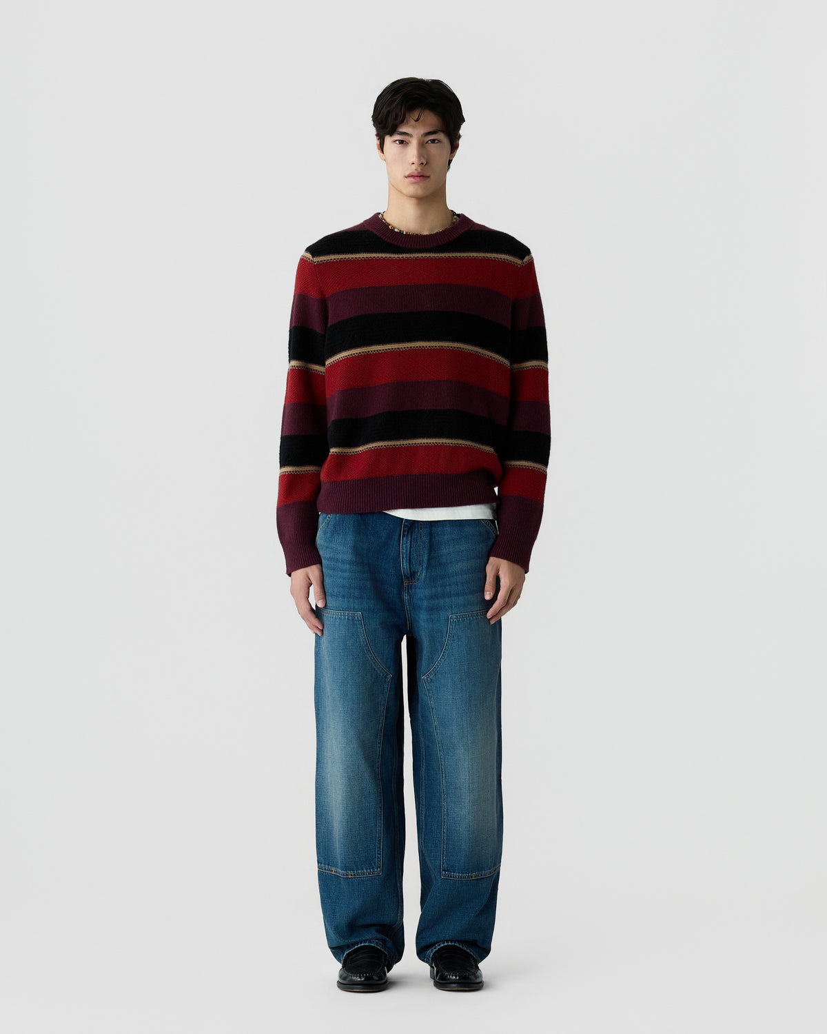 A young man stands against a plain background wearing a long-sleeve crewneck sweater with horizontal stripes in burgundy, black, and brown, paired with loose-fitting blue jeans and black shoes.