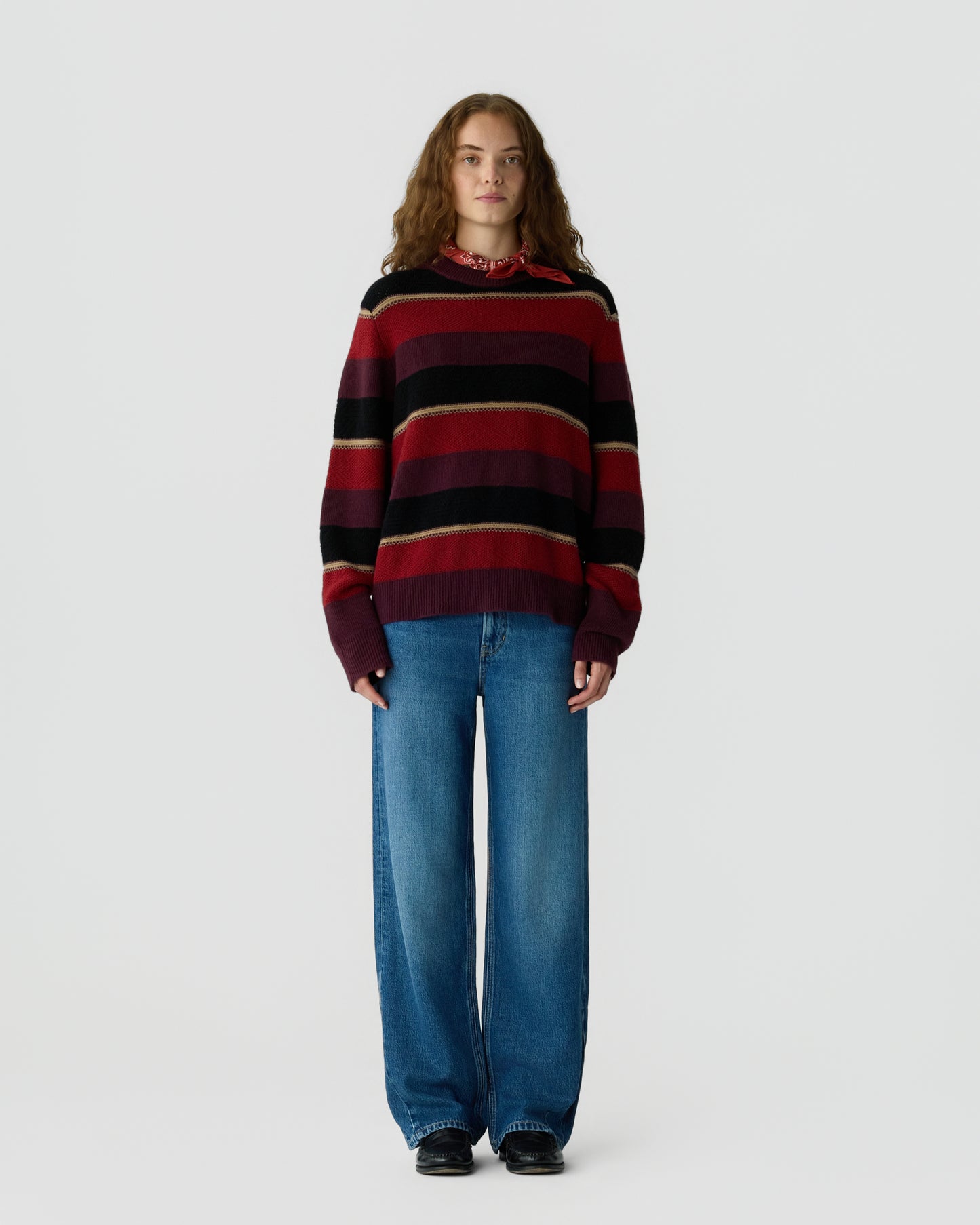 Young woman wearing a red, black, and beige striped sweater over a red patterned shirt, paired with wide-leg blue jeans and black shoes, standing against a plain white background.