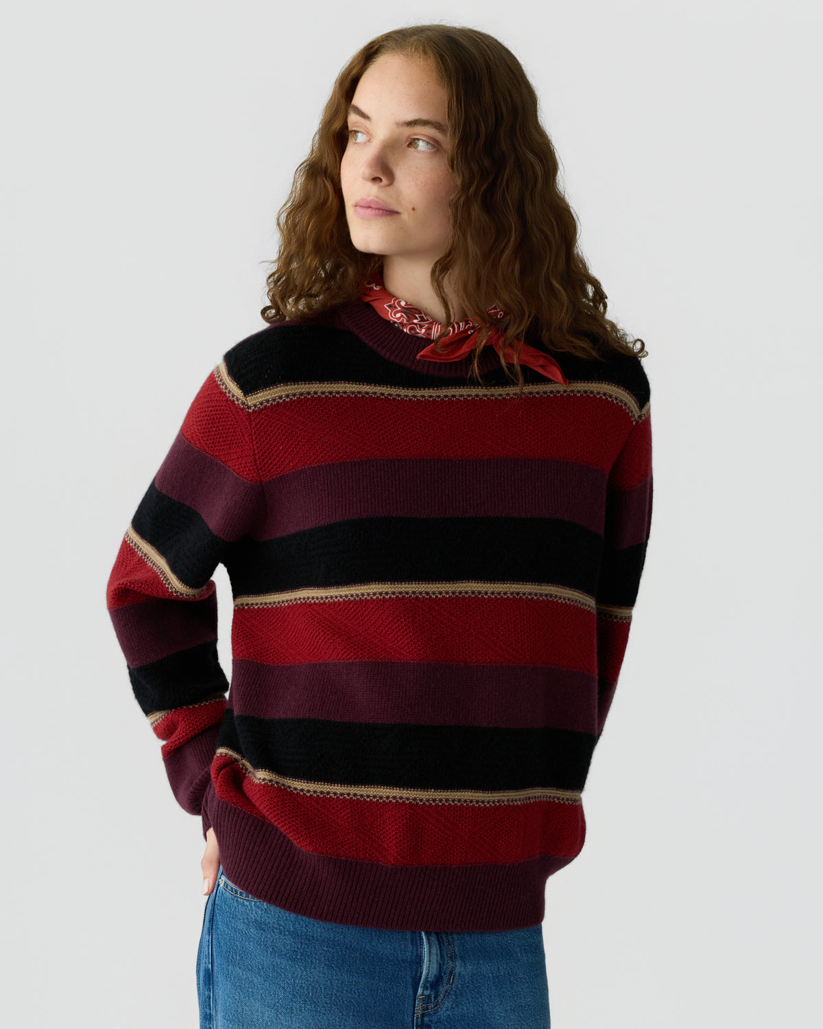 Young woman with curly hair wearing a red, black, and burgundy striped sweater and a red bandana around her neck, standing against a plain light background.
