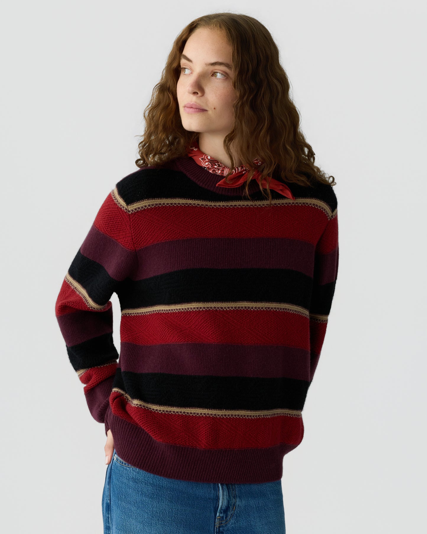 Young woman with curly hair wearing a red, black, and burgundy striped sweater and a red bandana around her neck, standing against a plain light background.