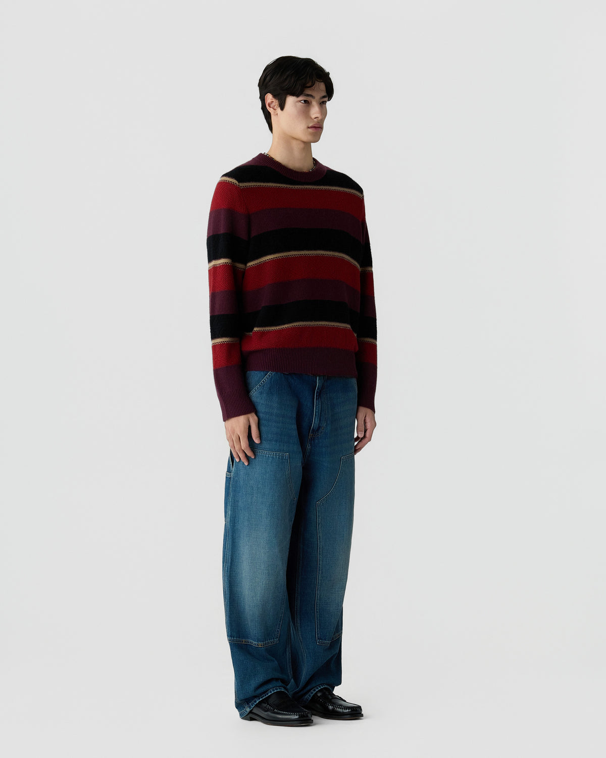 A man standing against a plain light background wearing a long-sleeve striped sweater in dark red, black, and beige paired with loose-fitting blue jeans and black shoes.