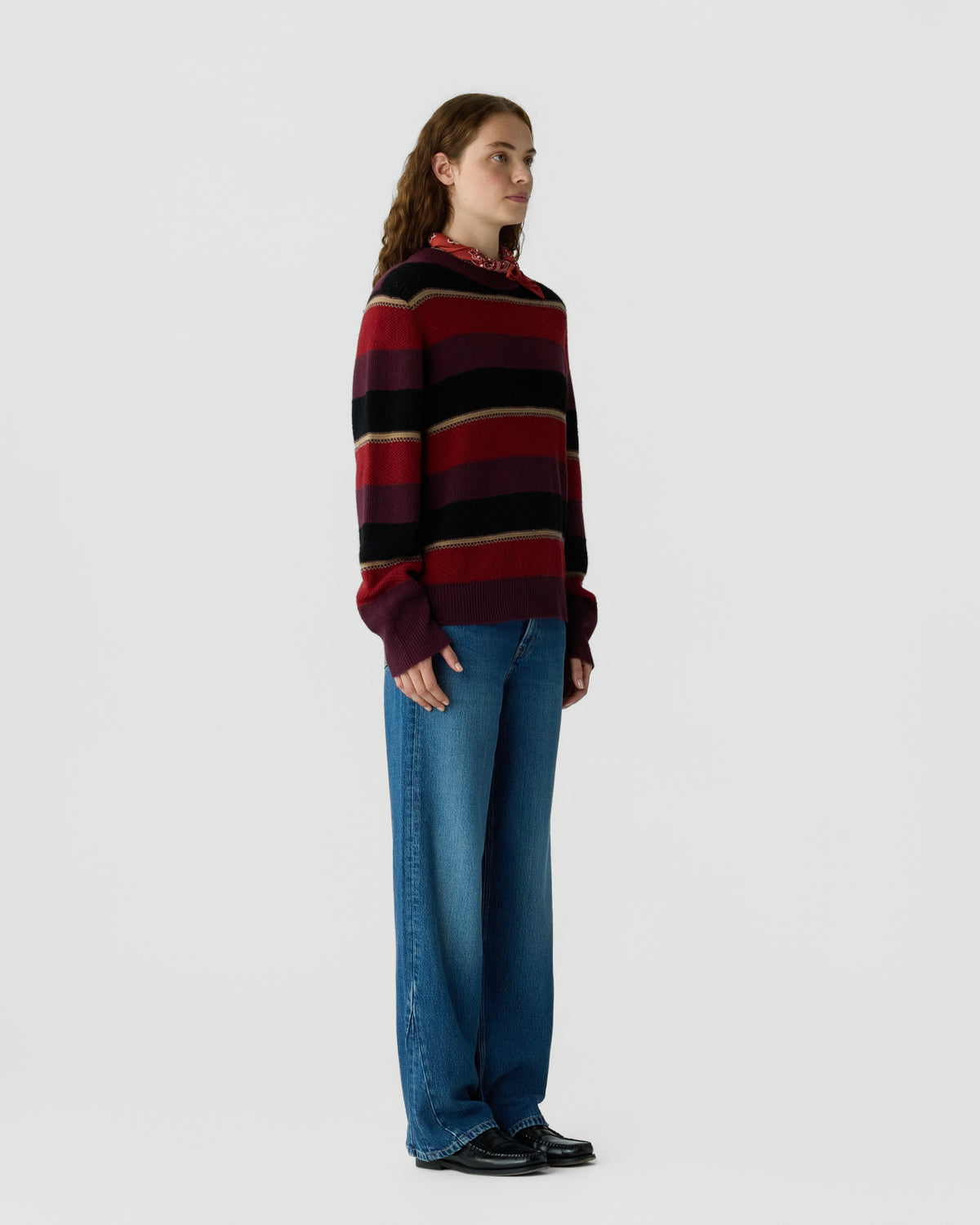 Woman standing against a plain background wearing a long-sleeve striped sweater in shades of red, black, and beige, paired with blue wide-leg jeans and black shoes.
