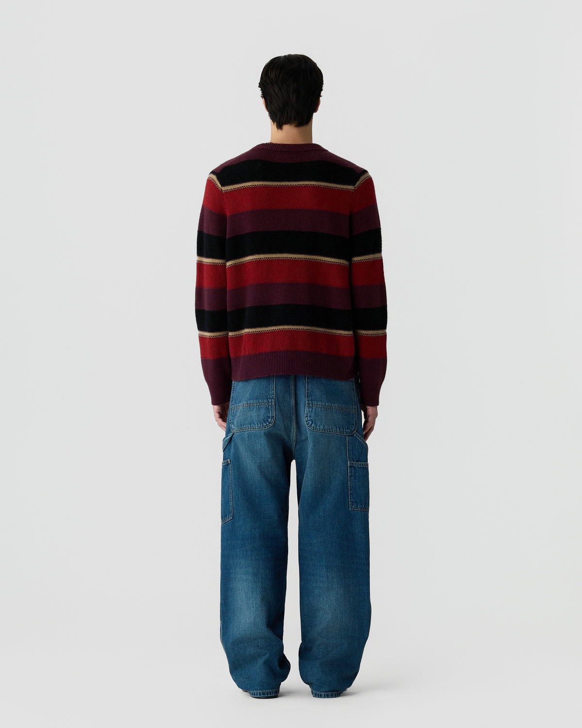 Man wearing a red, black, and beige striped sweater paired with loose-fitting blue denim jeans, shown from the back against a plain light background.