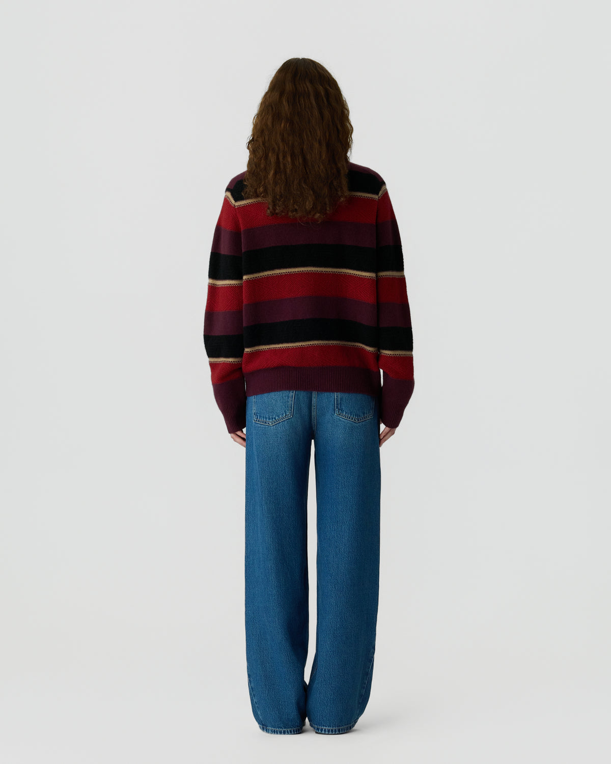 Person with long curly hair wearing a striped sweater in shades of red, black, and beige, paired with loose blue jeans, standing facing away against a plain background.