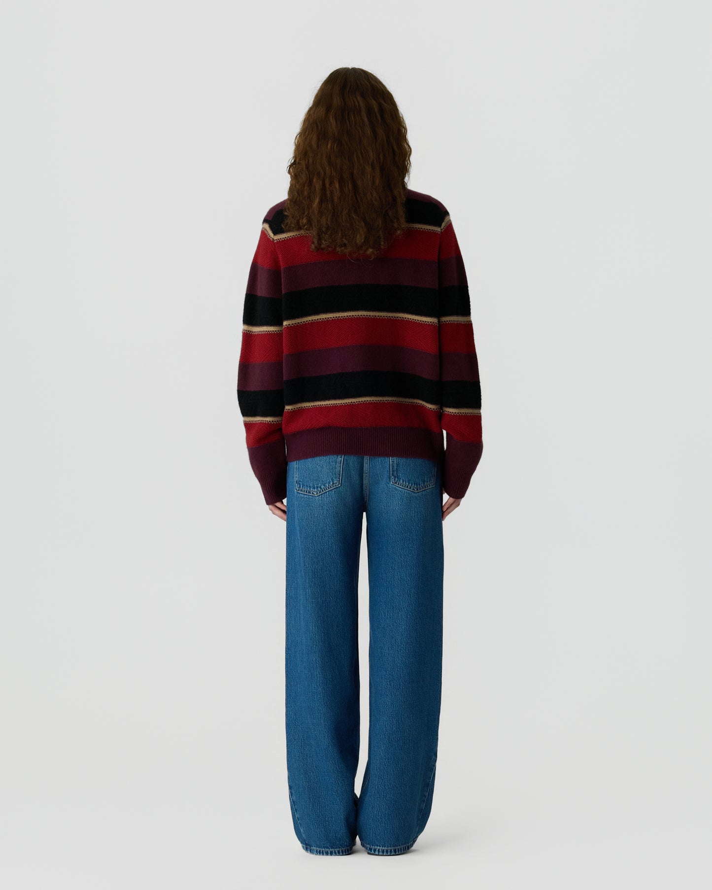 Person with long curly hair wearing a striped sweater in shades of red, black, and beige, paired with loose blue jeans, standing facing away against a plain background.