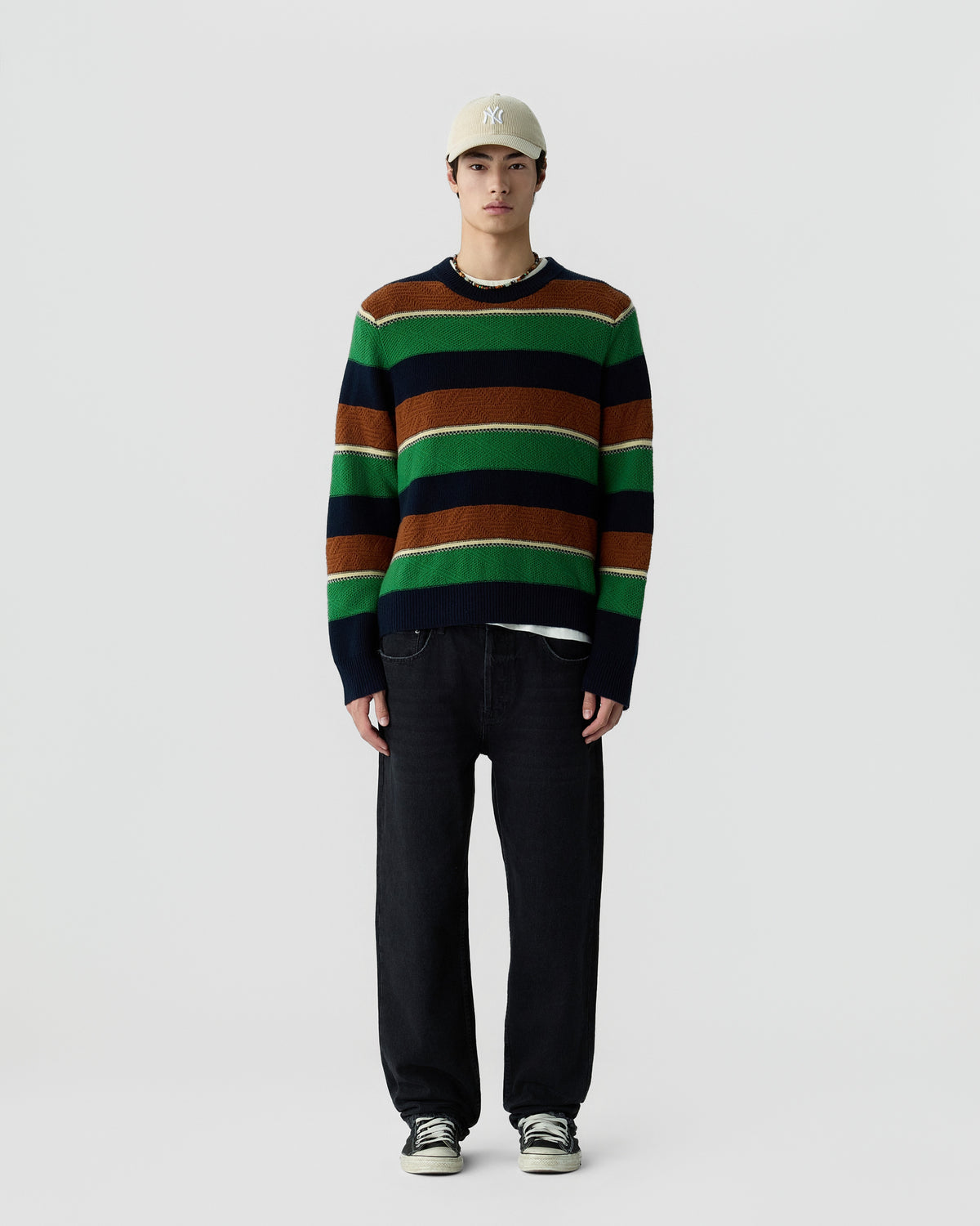 A young man standing against a plain light background wearing a green, brown, navy, and cream striped sweater, black pants, sneakers, and a beige baseball cap.