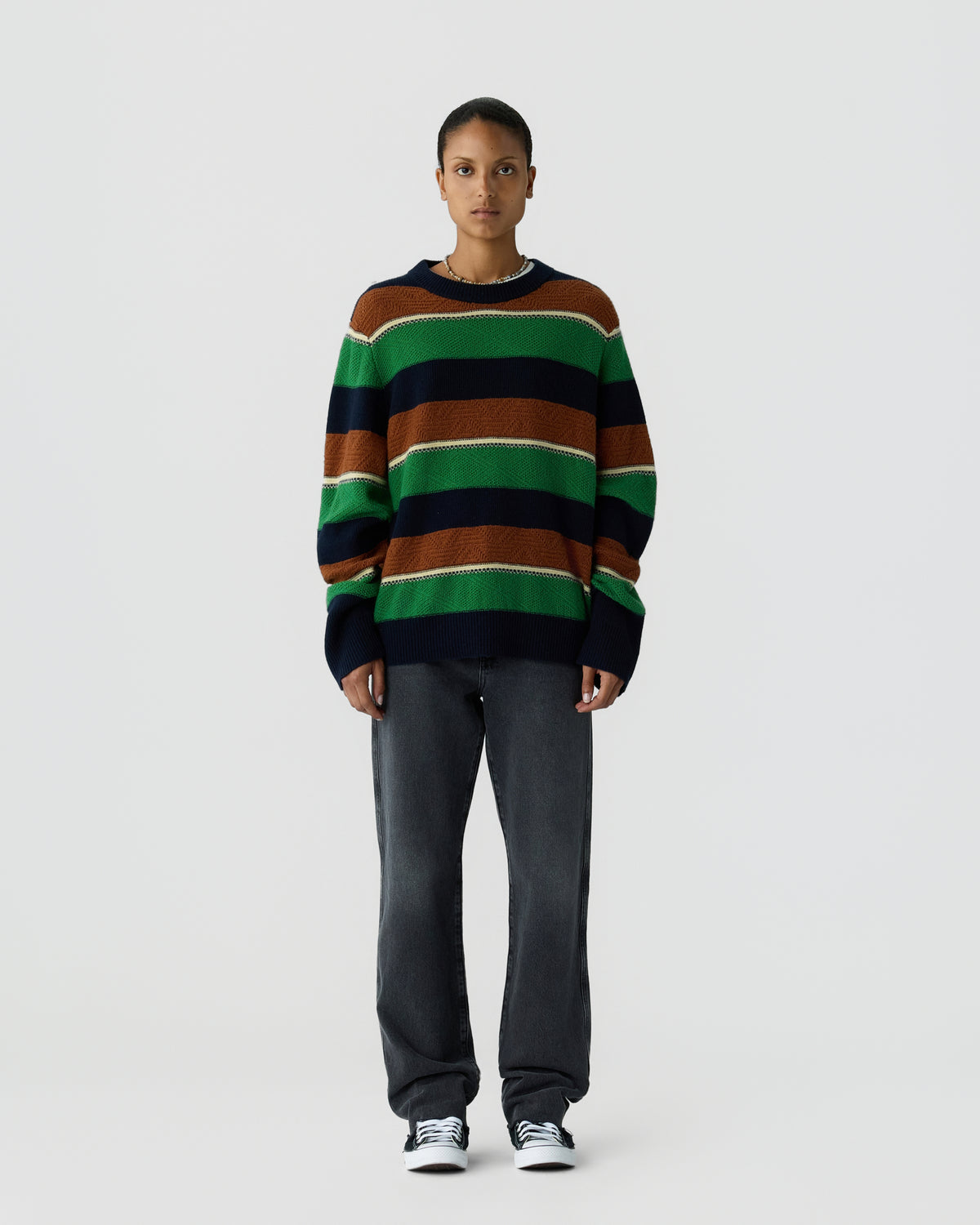 A person stands against a plain, light gray background wearing a loose-fitting striped sweater in green, brown, navy, and white, paired with dark gray pants and black and white sneakers.