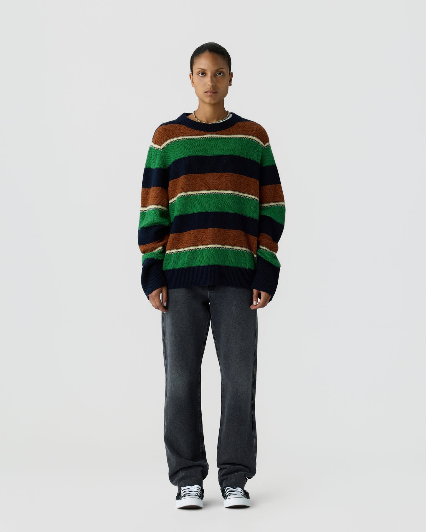 A person stands against a plain, light gray background wearing a loose-fitting striped sweater in green, brown, navy, and white, paired with dark gray pants and black and white sneakers.