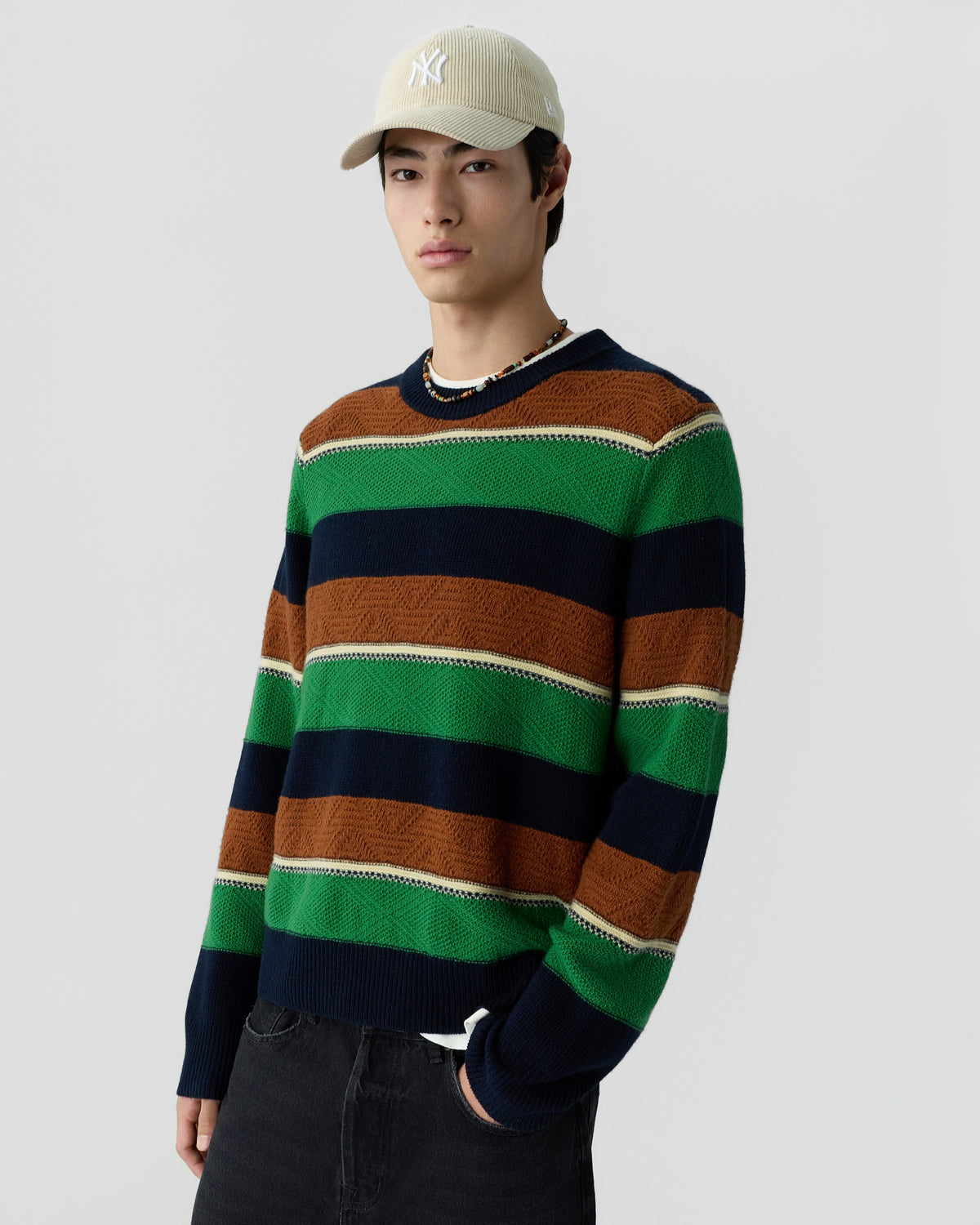 Young man wearing a striped sweater in green, navy, brown, and beige, paired with a beige baseball cap and dark pants against a plain light gray background.