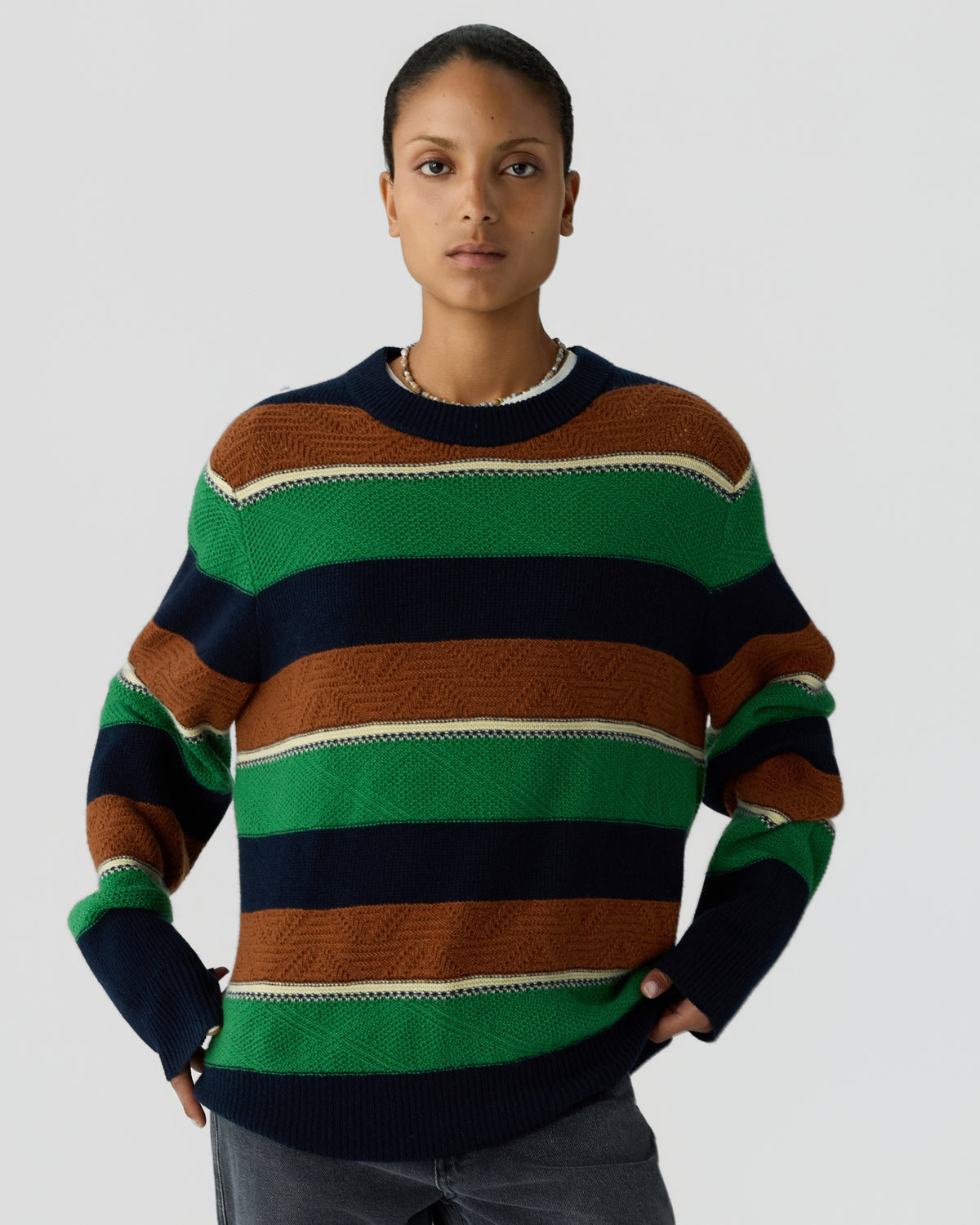 Woman wearing a long-sleeve crewneck sweater with wide horizontal stripes in green, navy blue, brown, and beige, standing with hands in pockets against a plain background.