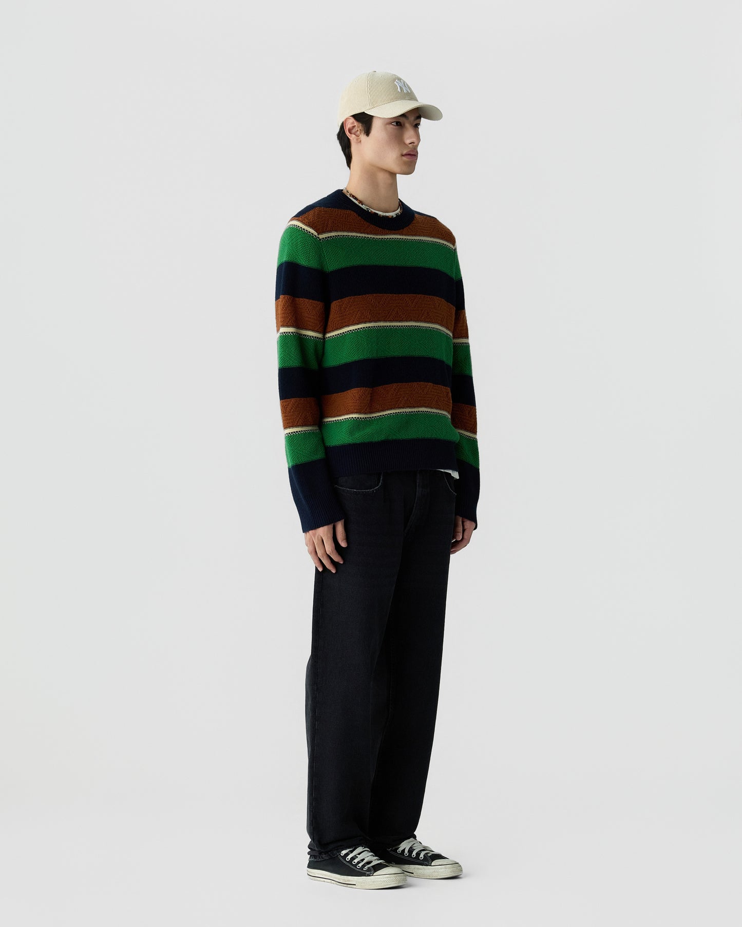 A man wearing a beige baseball cap, a green, brown, and navy striped sweater, dark pants, and black and white sneakers standing against a plain light gray background.