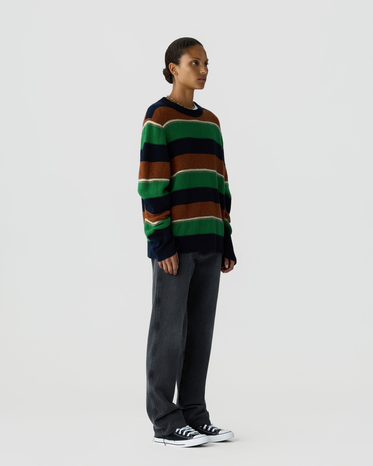 Side view of a person wearing a loose-fitting striped sweater with green, brown, and navy blue bands, paired with relaxed dark gray pants and black sneakers against a plain background.