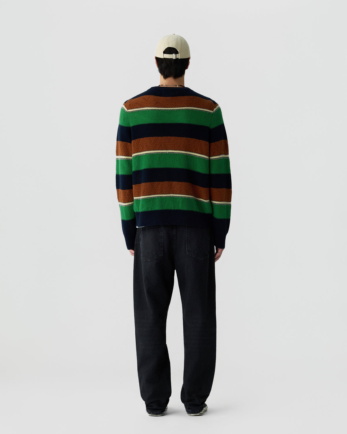 Person wearing a striped sweater in green, brown, navy, and white, paired with dark loose-fitting jeans and a light-colored baseball cap, shown from the back.