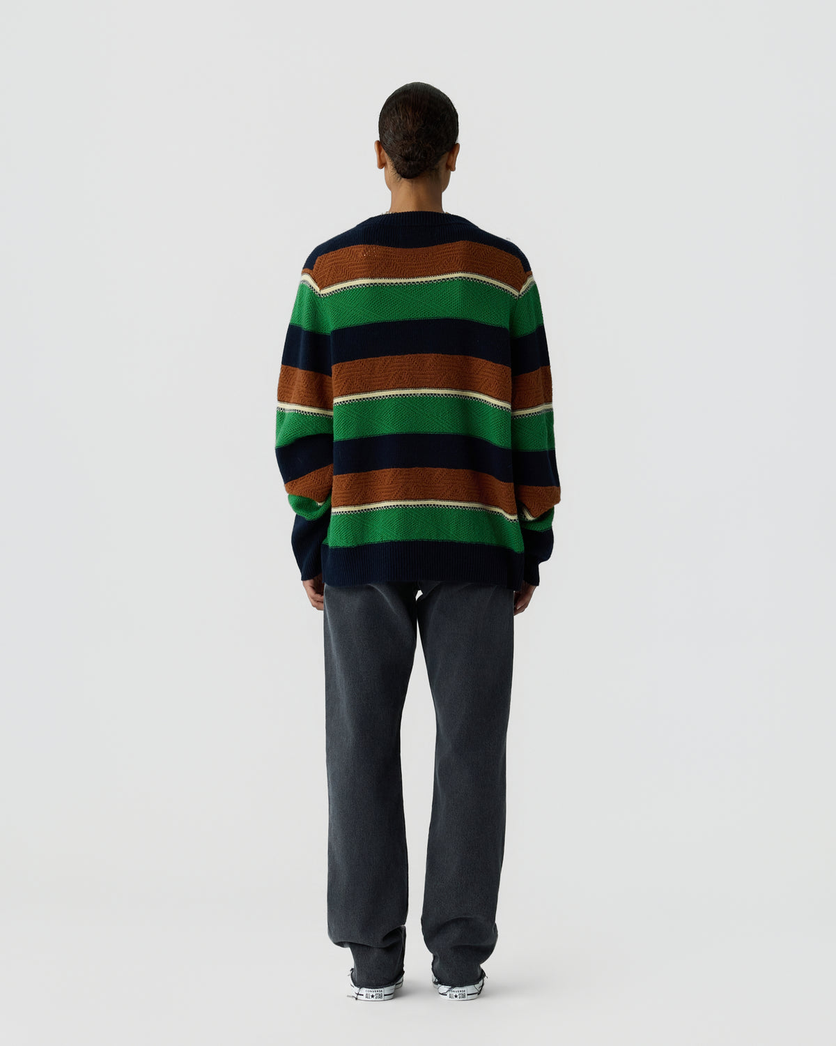 Person standing with back facing camera wearing a striped sweater in navy blue, green, and brown with dark gray pants and white sneakers against a plain white background.