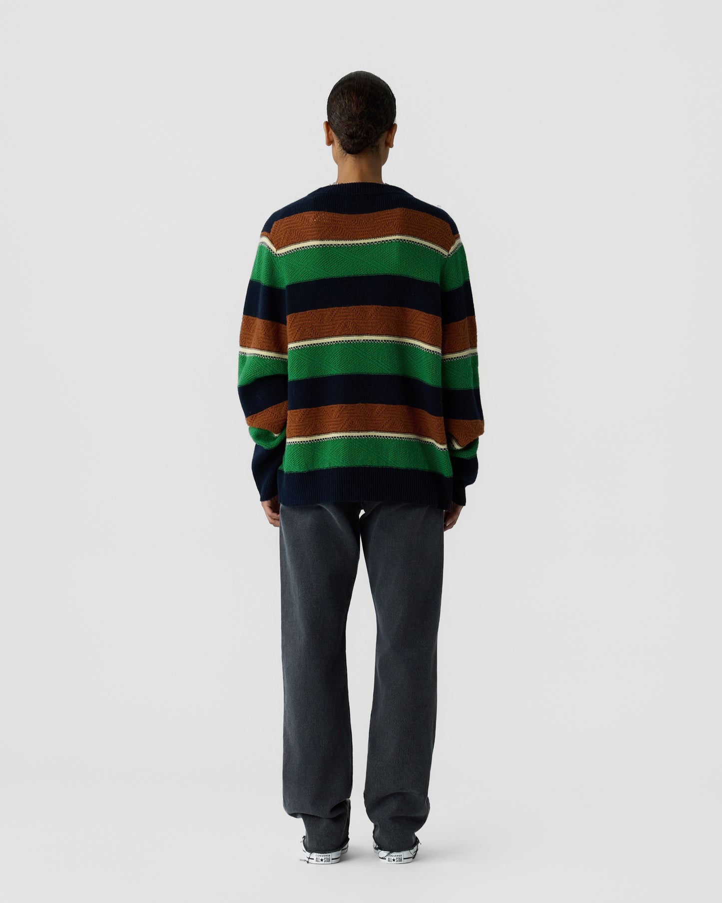 Person standing with back facing camera wearing a striped sweater in navy blue, green, and brown with dark gray pants and white sneakers against a plain white background.