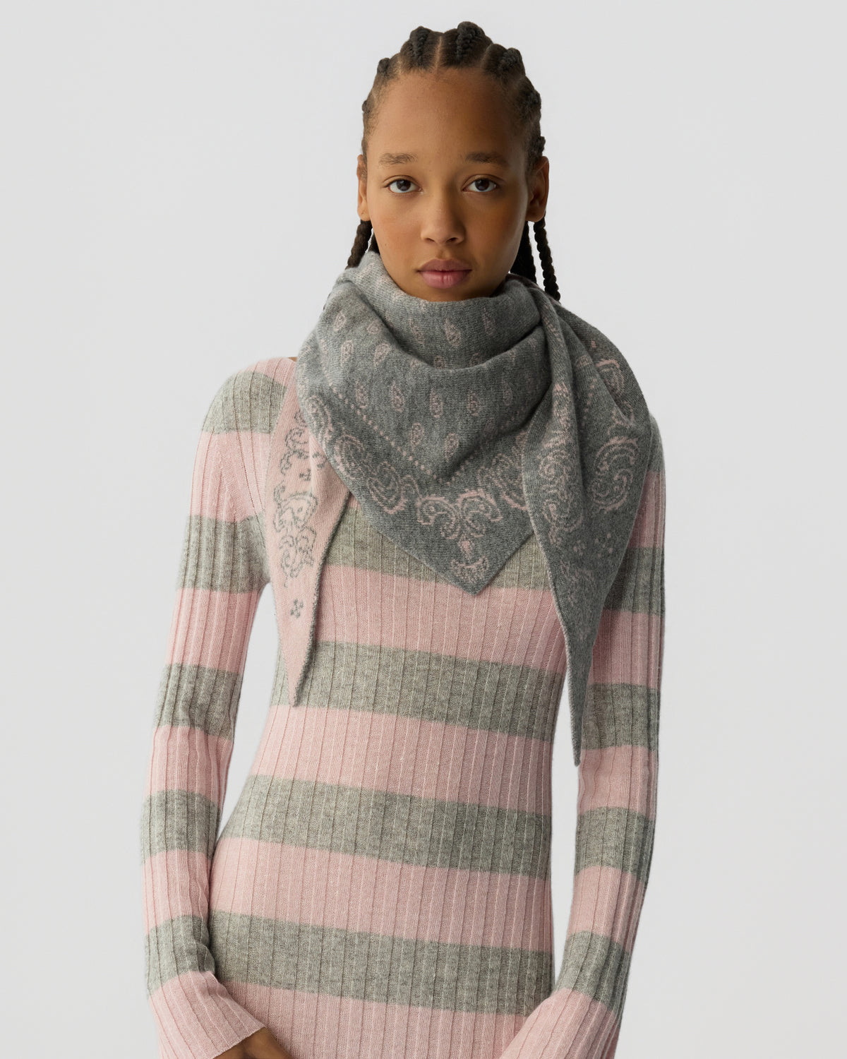 Young woman with braided hair wearing a ribbed knit dress featuring wide pink and grey stripes, paired with a large grey patterned scarf wrapped around her neck.