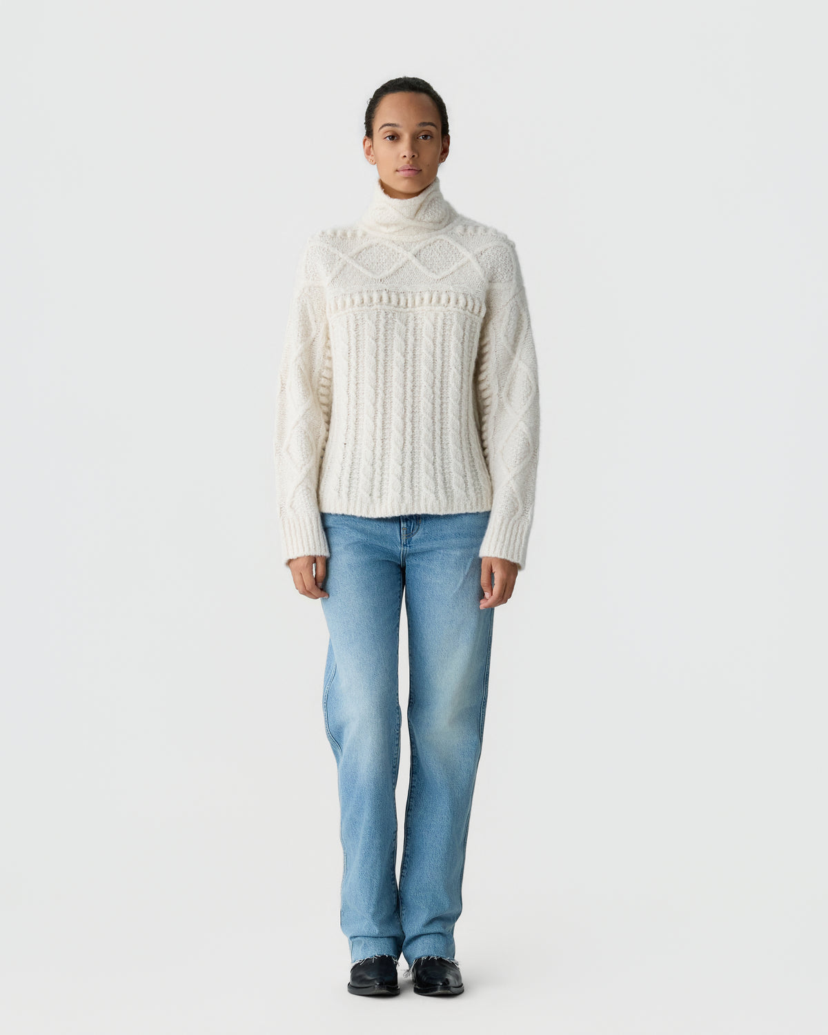 Person standing against a light gray background wearing a cream cable knit turtleneck sweater, blue jeans, and black shoes