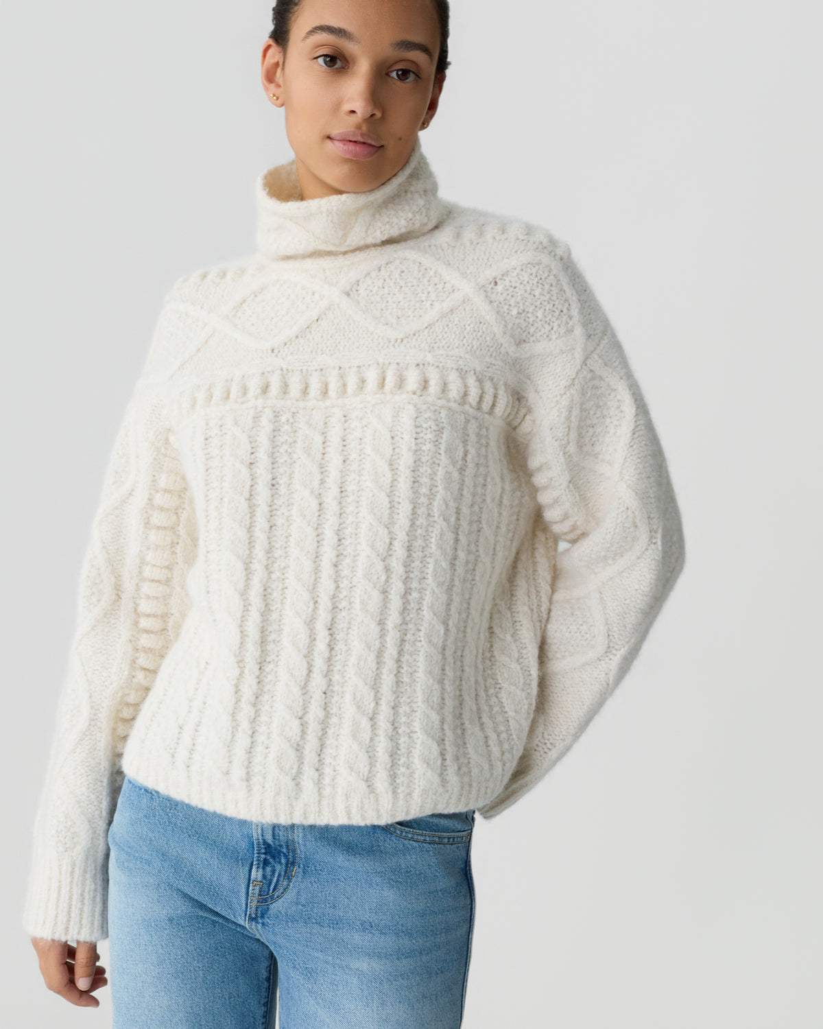 Person wearing a cream cable knit turtleneck sweater with textured patterns and blue jeans against a light gray background
