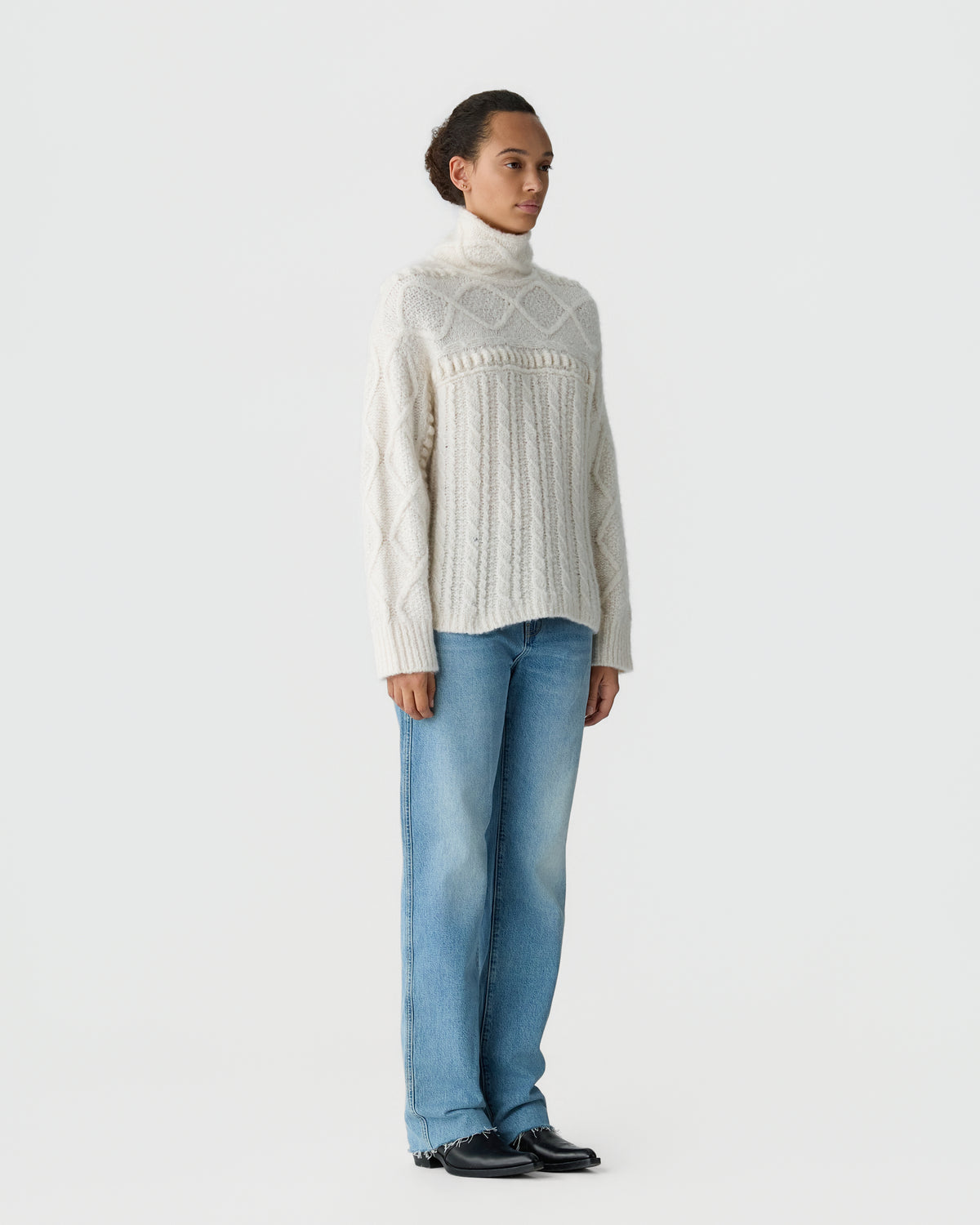 Woman wearing a cream cable knit turtleneck sweater with blue jeans and black shoes against a plain light gray background