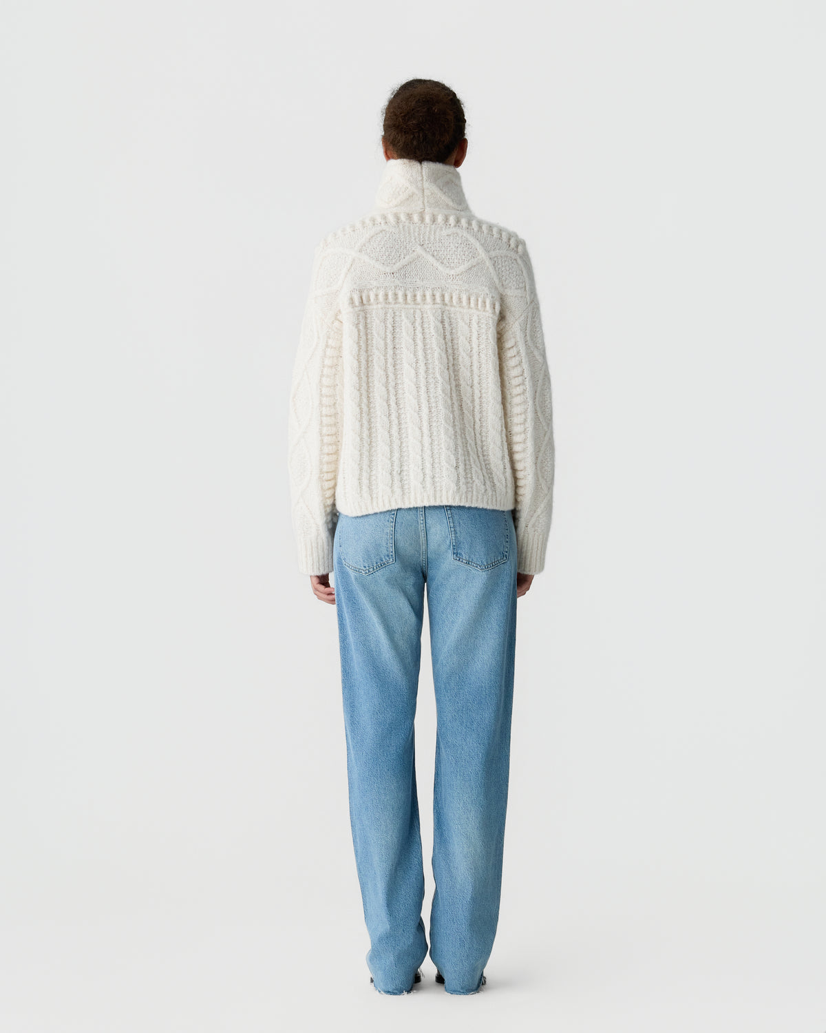 Back view of a person wearing a cream cable knit turtleneck sweater and light blue jeans against a plain light gray background