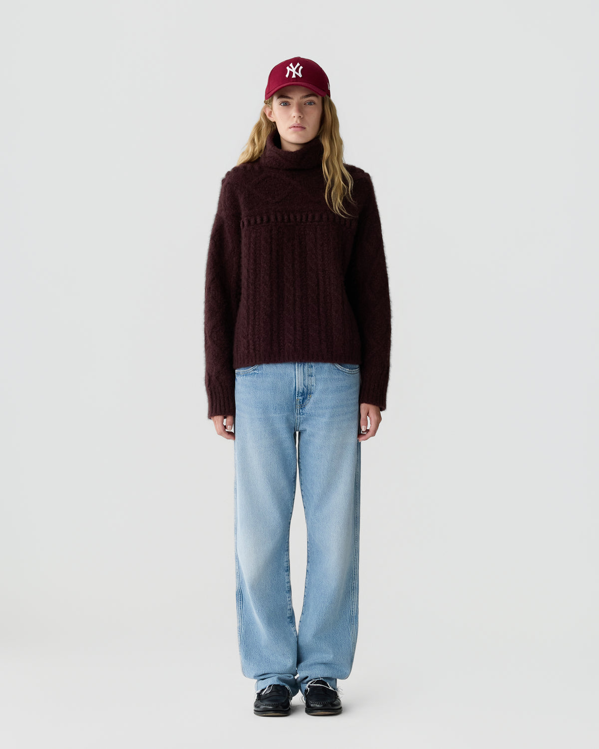 Model wearing a burgundy knitted turtleneck sweater with light blue jeans a red baseball cap and black shoes against a pale gray studio background