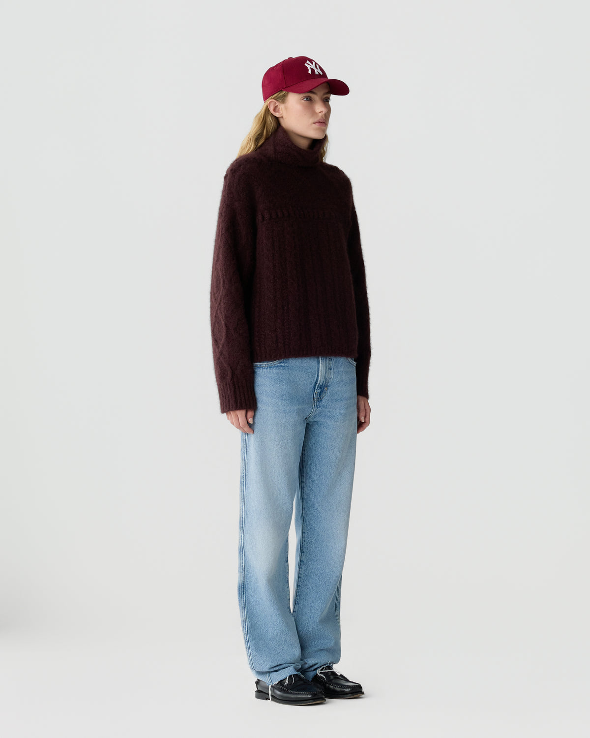 Woman wearing a burgundy knit sweater light blue wide leg jeans black shoes and a red baseball cap against a pale gray background