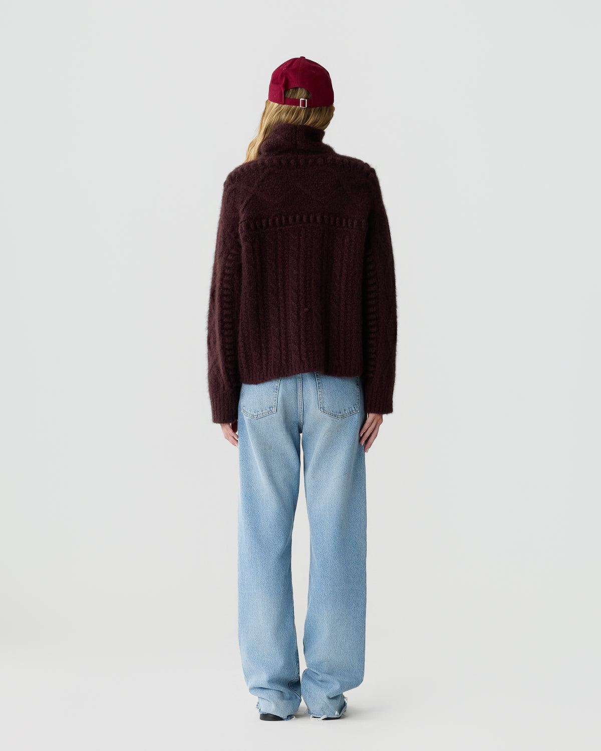 Person viewed from behind wearing a dark burgundy knit sweater light blue jeans and a red cap against a pale gray background