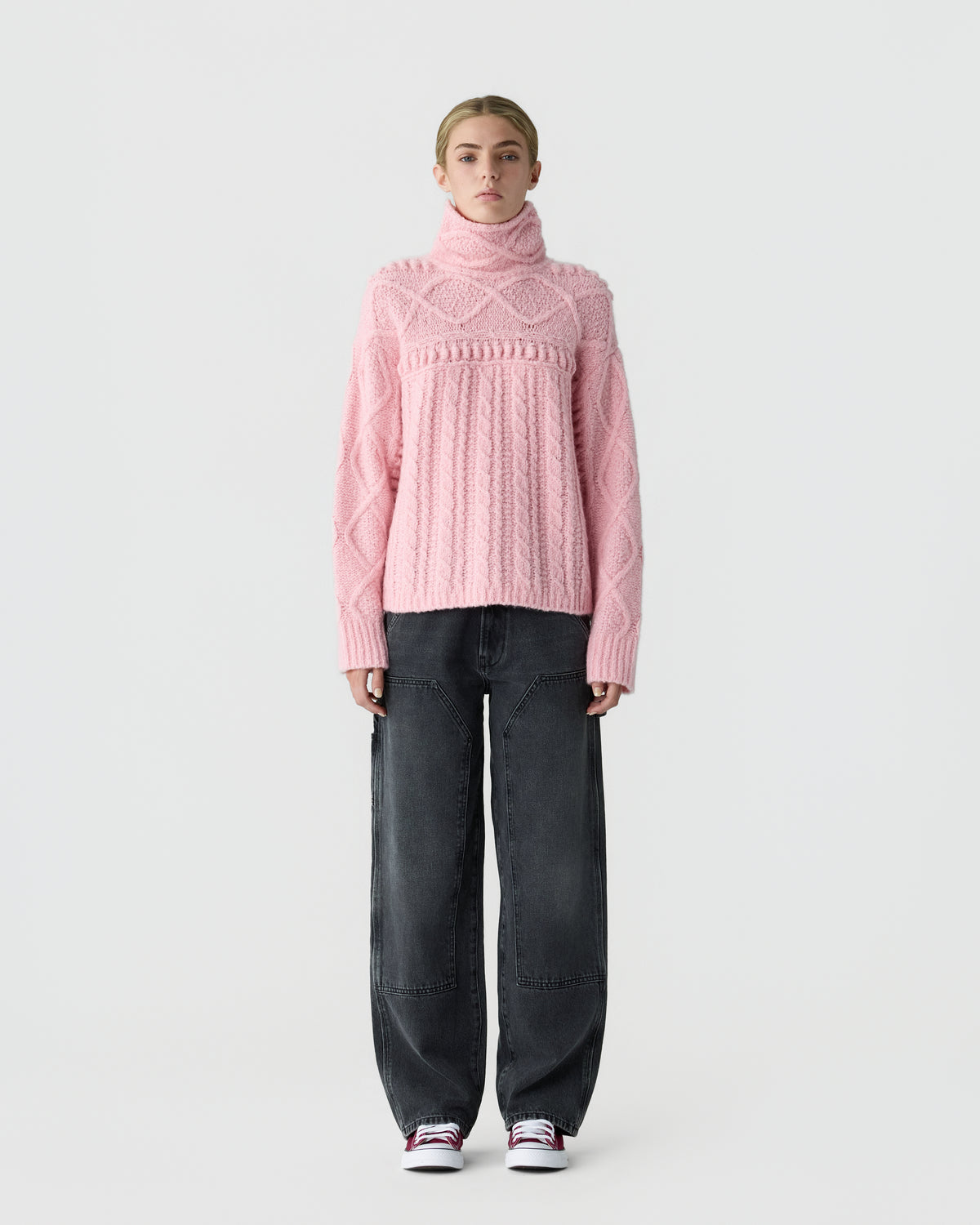 Model wearing a pink cable knit turtleneck sweater with black wide leg jeans and white sneakers against a light gray background