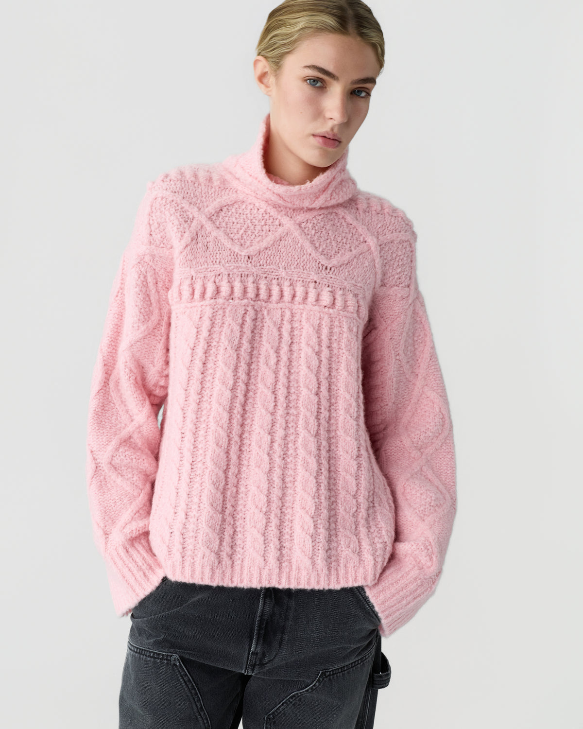 Woman wearing a soft pink textured turtleneck sweater with cable knit detailing and black jeans against a light gray background